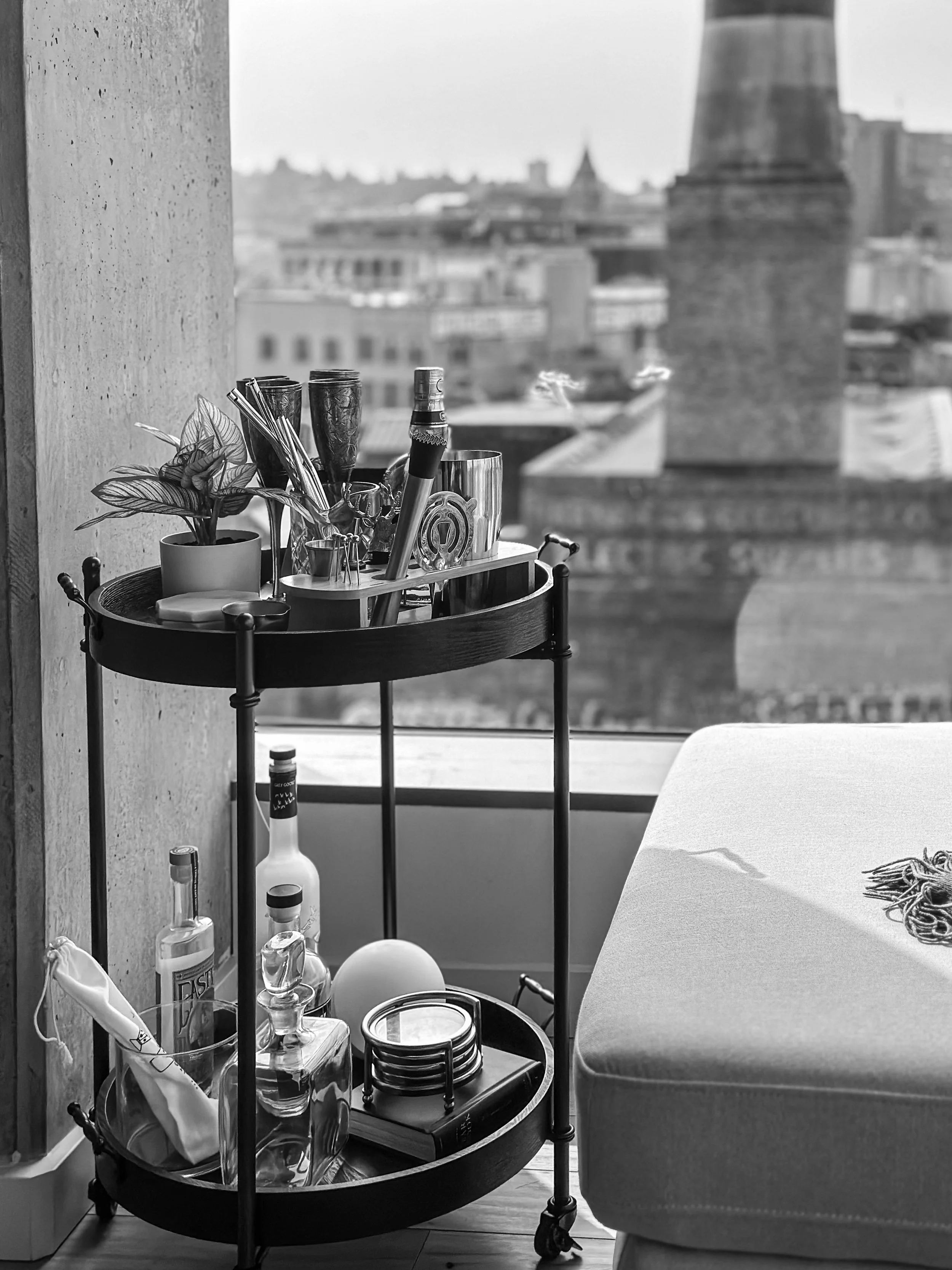 A black and white photo of a round bar cart with various bottles, glasses, and a potted plant near a large window with city buildings in the background.