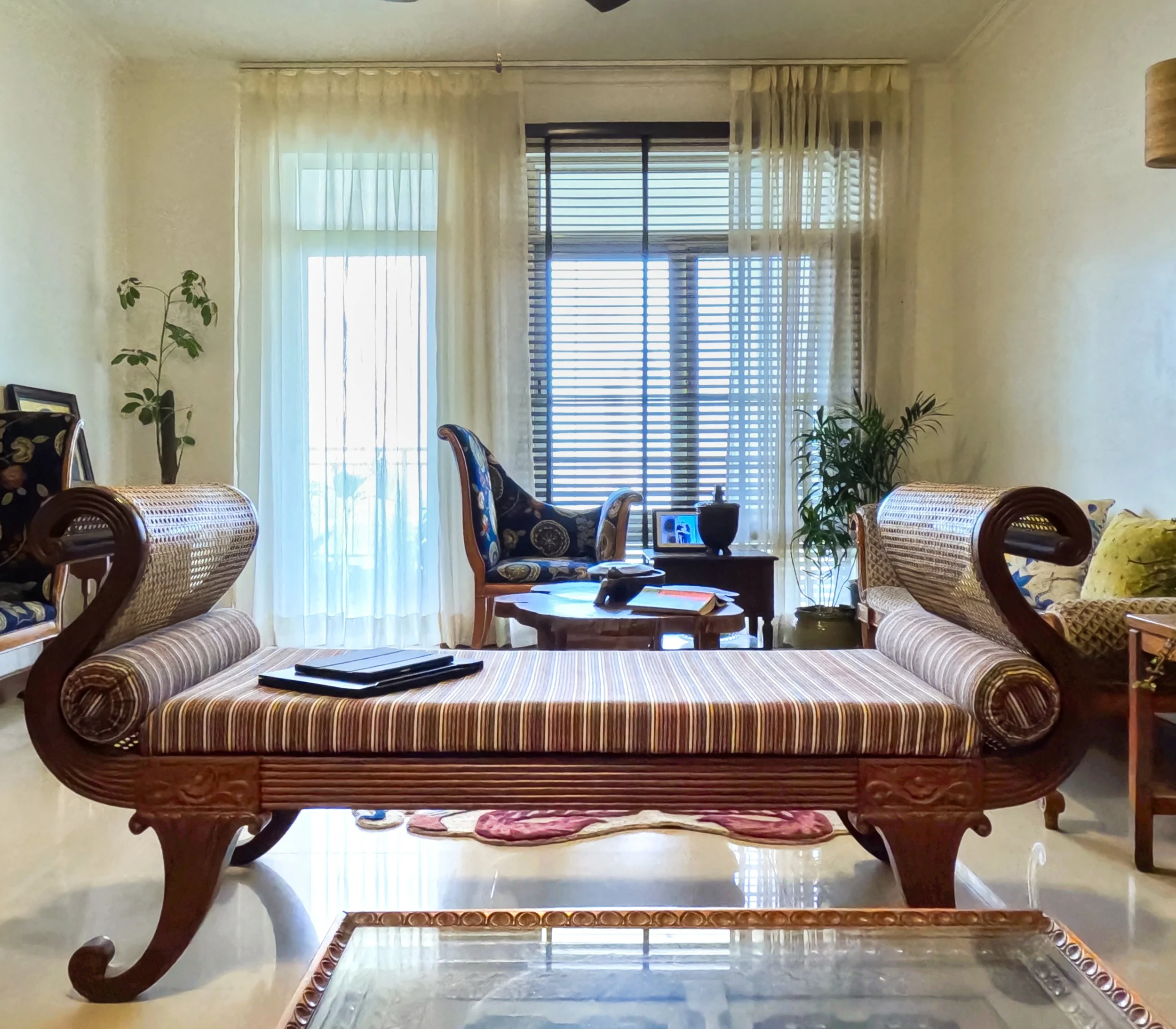 Living room with antique-style wooden chaise lounge, plaid upholstery, and nearby armchair with floral fabric, multiple windows with sheer curtains, and indoor plants.