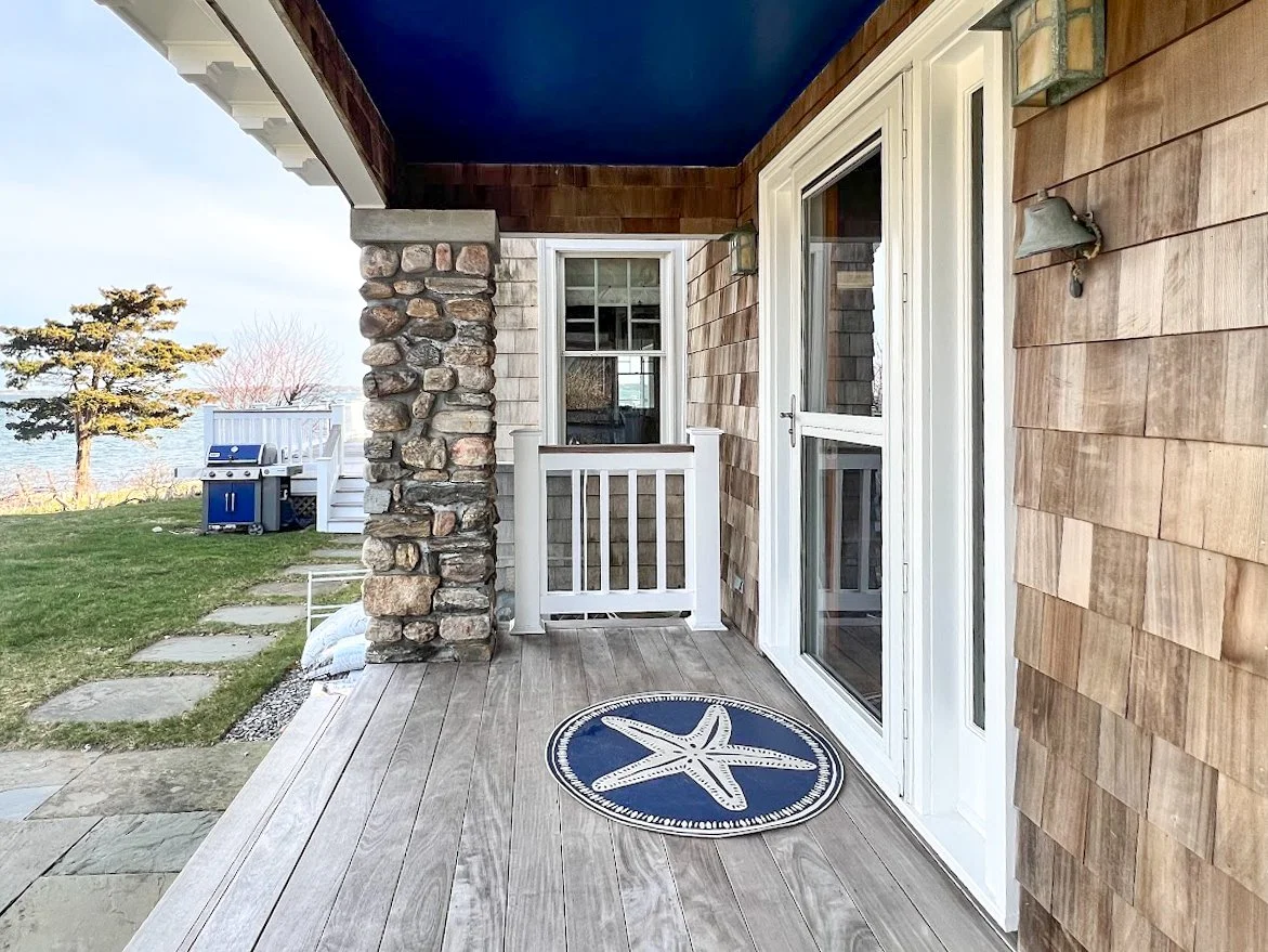 A front porch with wooden flooring, a starfish-themed rug, a white door, and sliding glass doors. The porch has a stone pillar and a wall-mounted light fixture. In the background, there is a grassy yard with stepping stones, a tree, and a blue grill