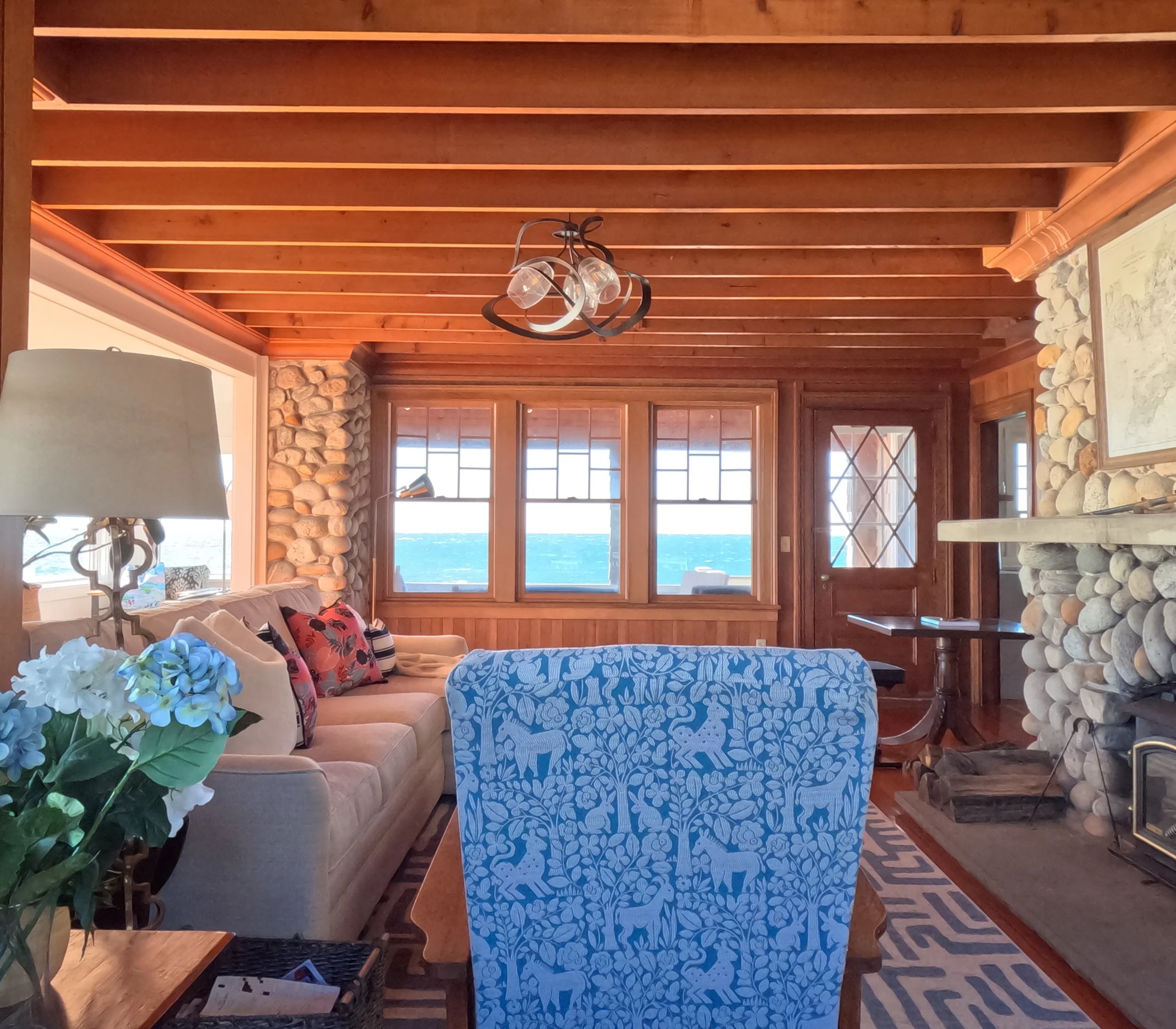 Interior of a cozy living room with wooden ceiling and stone fireplace, beige sofa with colorful pillows, blue armchair with white animal and plant pattern, windows with ocean view, and decorative lamps and artwork.
