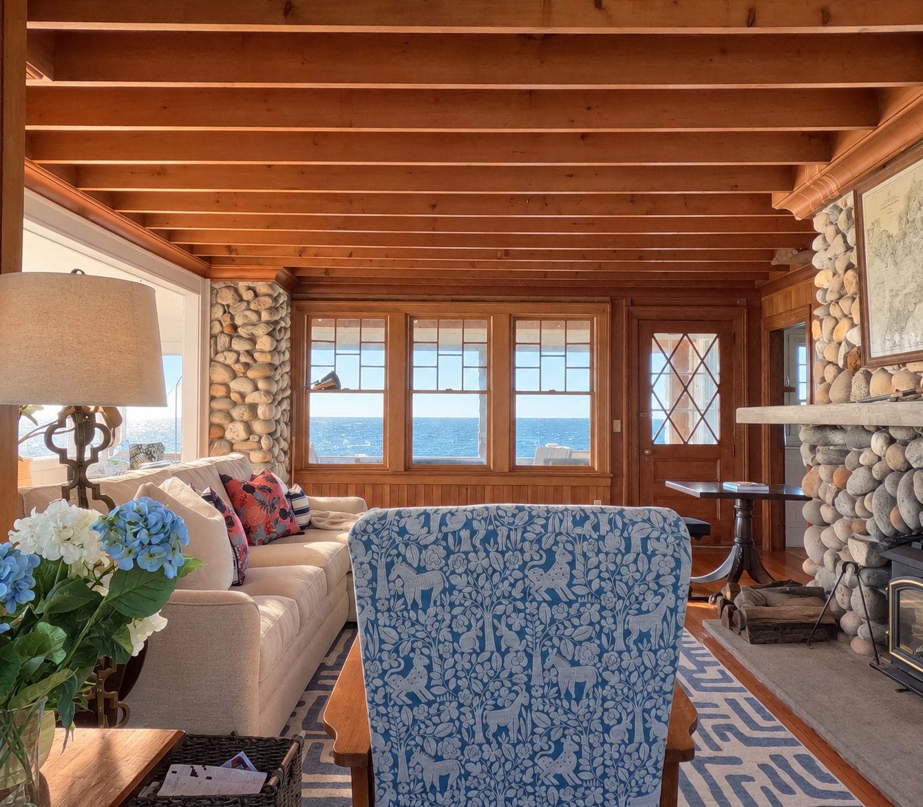 A cozy living room with large windows showing an ocean view, featuring a beige sofa with patterned pillows, a blue and white patterned armchair, a stone fireplace, a wooden ceiling, and a table with a book on it.