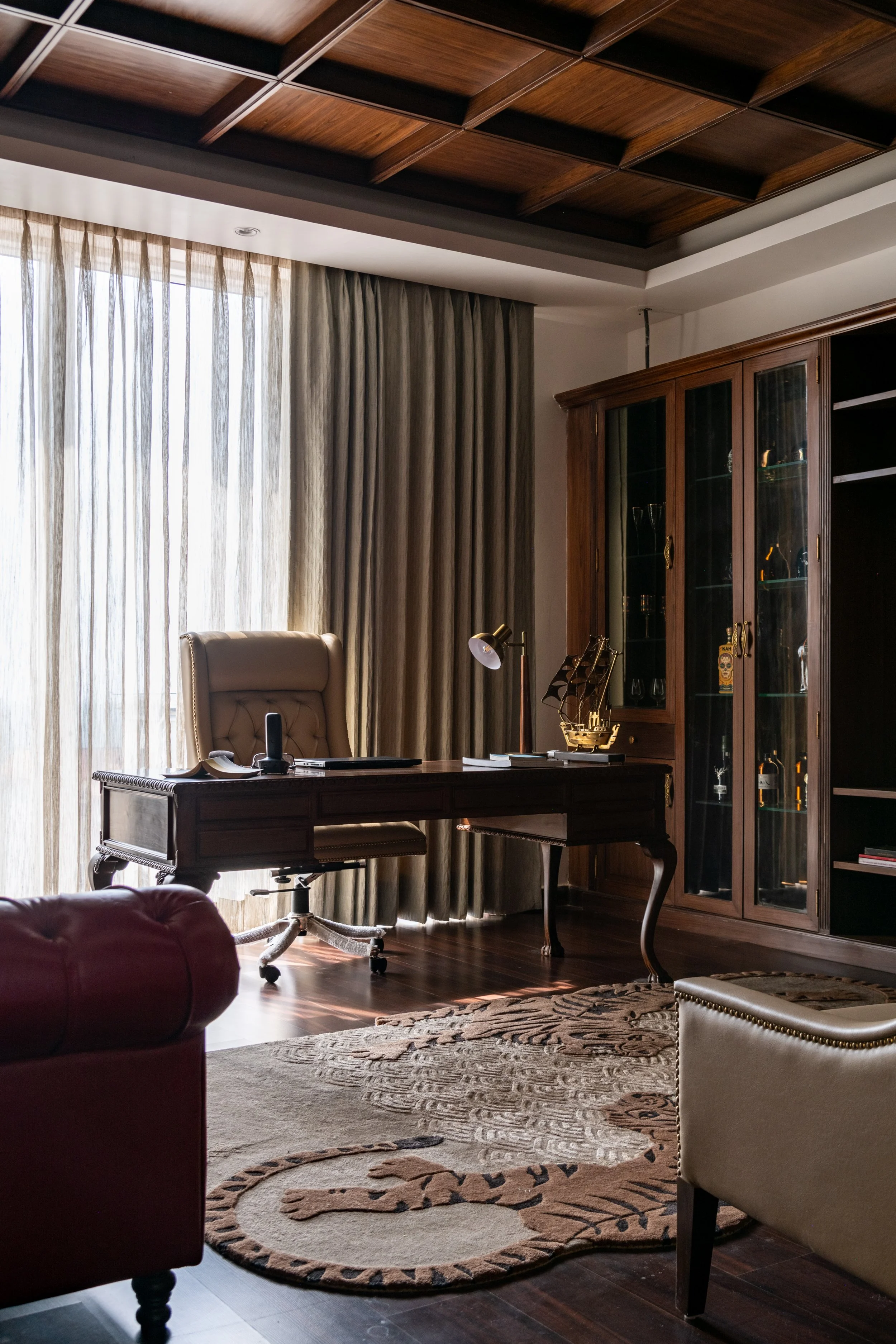 Home office with a large wooden desk, beige leather office chair, bookshelf with glass doors, and a decorative rug on wooden floor near a window with sheer curtains.