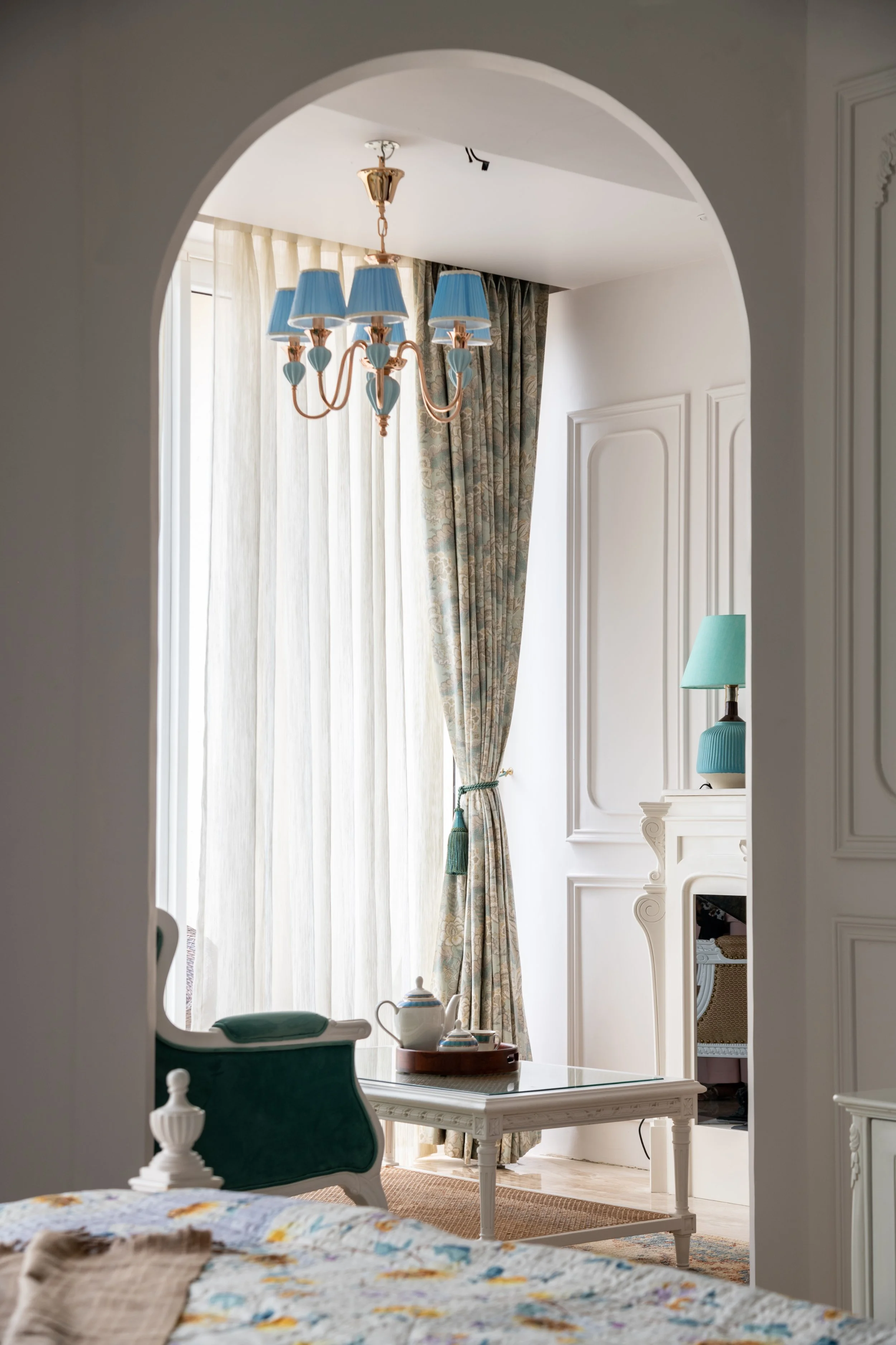 A room viewed through an arched doorway, featuring a chandelier with blue lampshades, patterned curtains, a white fireplace with a blue lamp, a small white table with a teapot, and a green and white armchair.