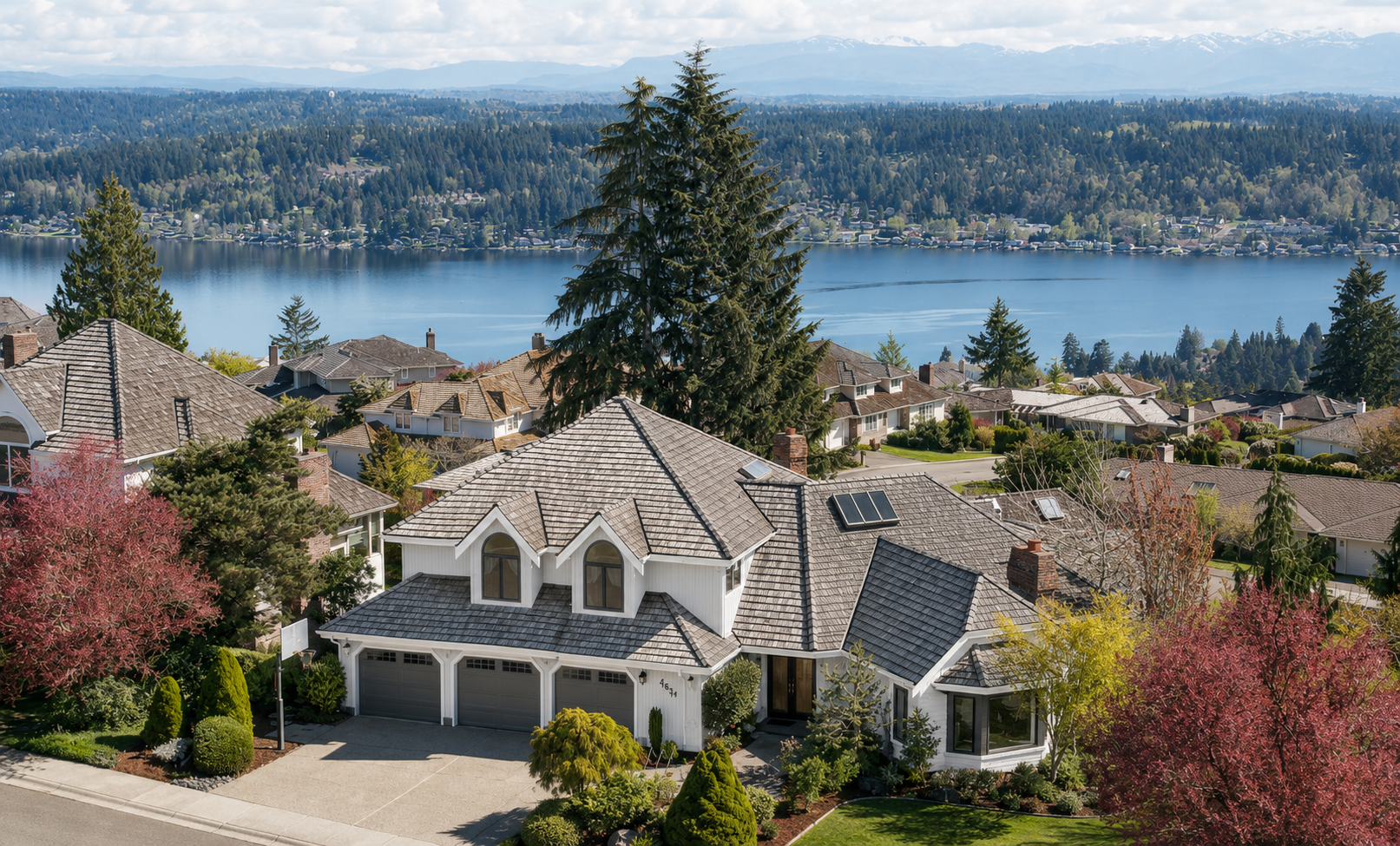 Aerial view of a white house with a brown roof, surrounded by trees, with a lake and forested hills in the background.