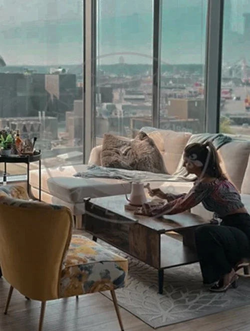 A woman wearing a mask working at a desk in a modern living room with large windows, cityscape view, beige sofa with stuffed animal, and a yellow armchair.