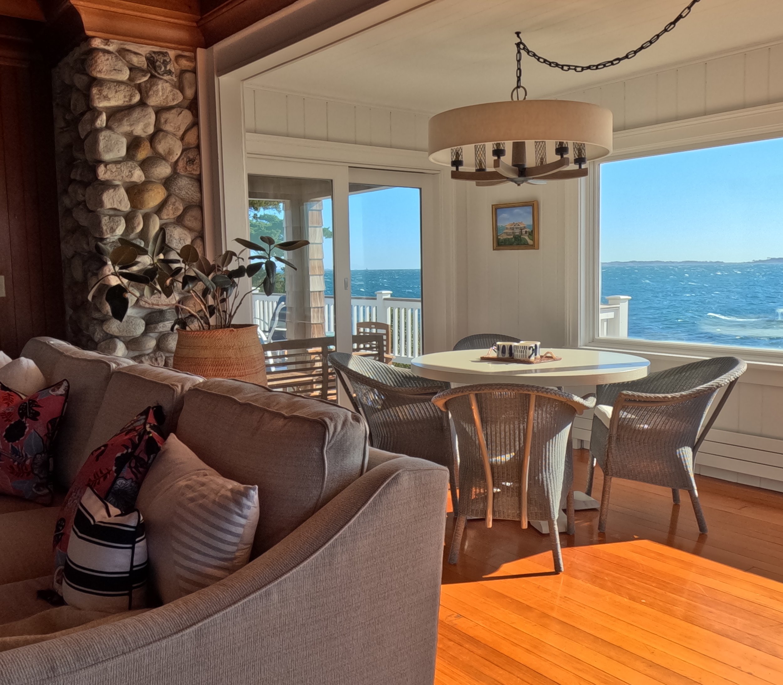 Living room with a view of the ocean, featuring a beige sofa with patterned pillows, a white round dining table with wicker chairs, and a large window showing the water and blue sky.