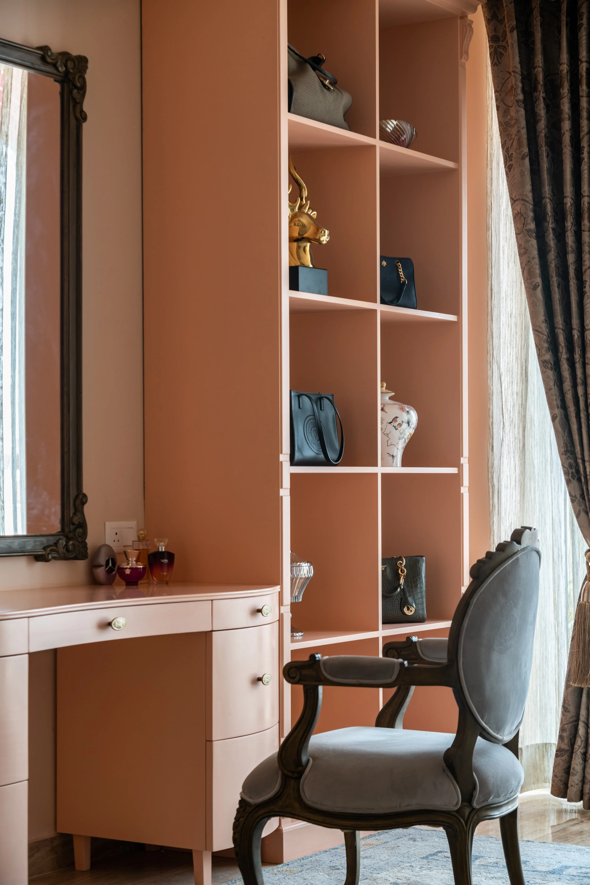 A pink shelving unit with handbags and decorative items, a gray armchair in front, beside a window with curtains, and a pink vanity table with perfume bottles and a mirror.