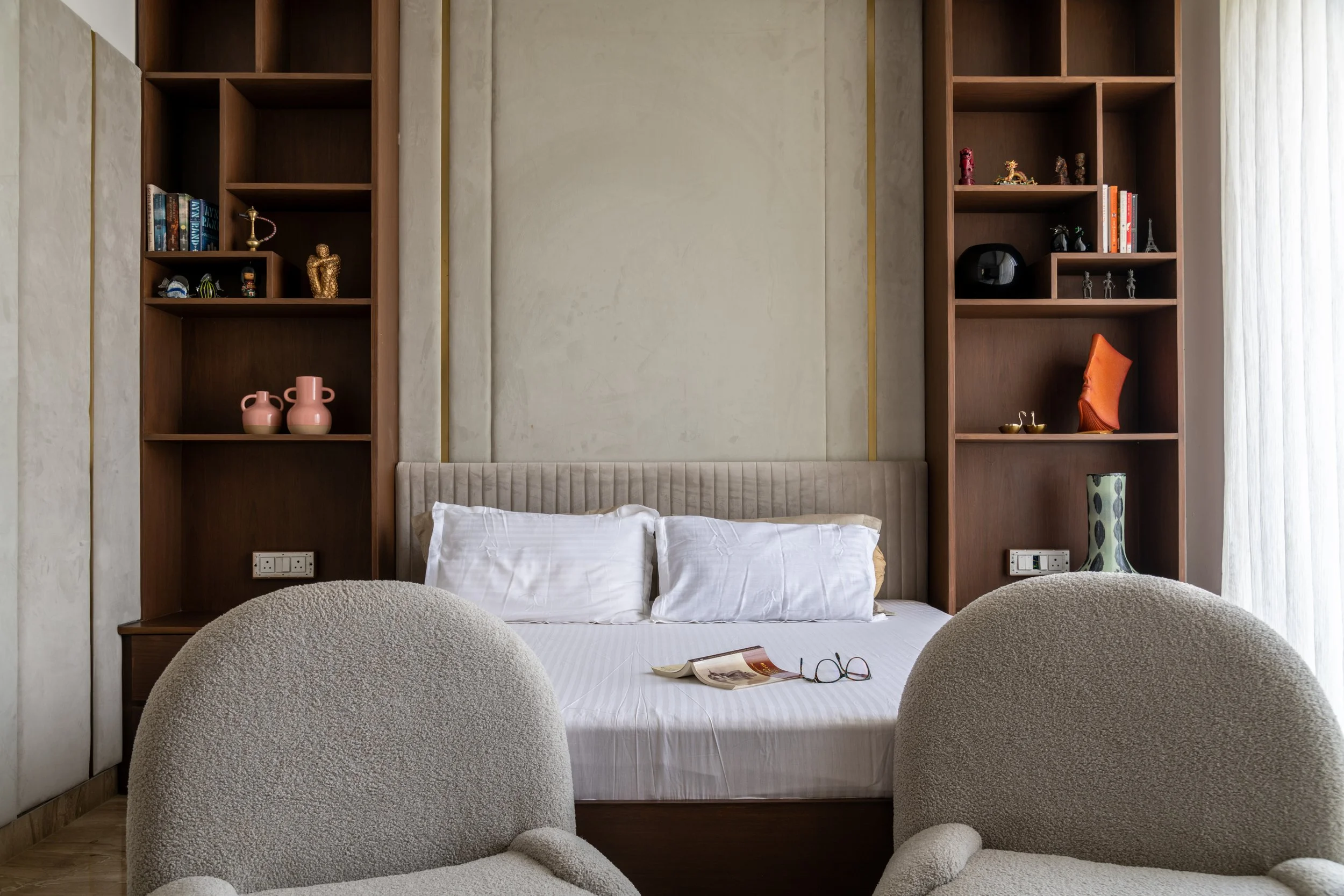 A bedroom with a bed, two white pillows, a pair of glasses, an open magazine, and two beige armchairs. Wooden built-in shelves with decorative items and books are on either side of the bed, and a window with sheer white curtains is on the right.