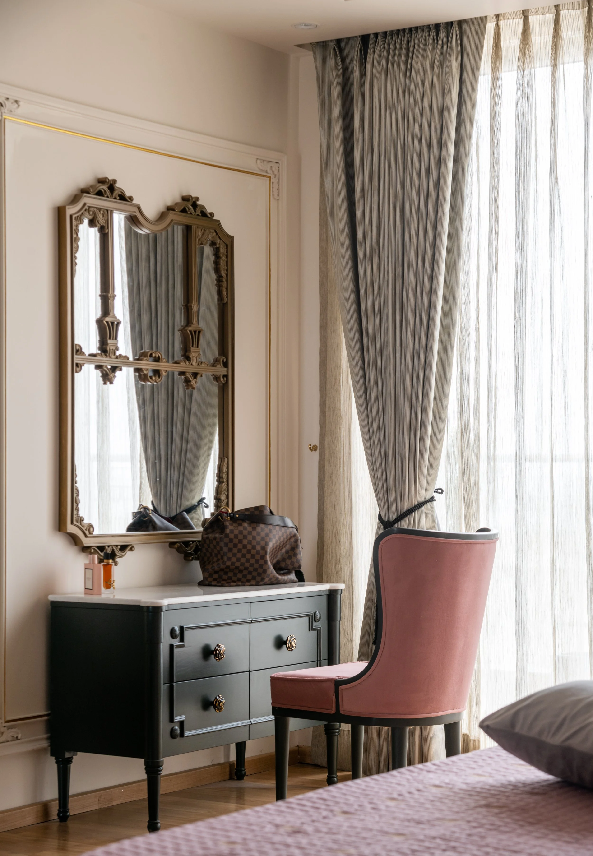 A bedroom corner with a black dresser, a pink upholstered chair, a mirror, curtains, and a tote bag on the dresser.