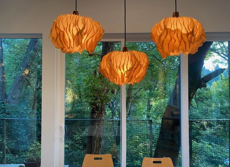 Three orange pendant lights hanging above a table in front of large glass windows showing trees outside.