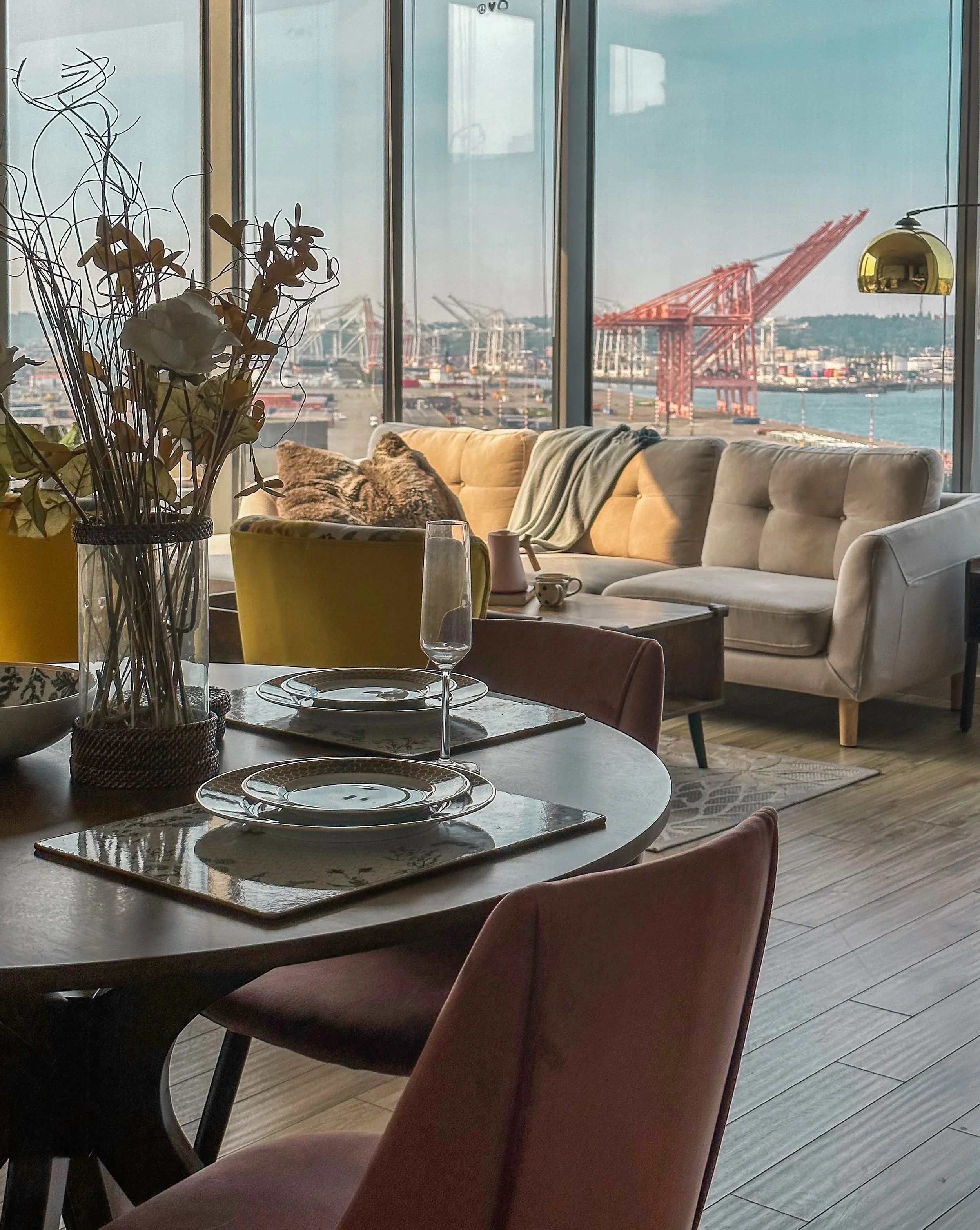 Interior of a modern apartment with a view of a harbor and large red cranes outside the glass windows, decorated with a dining table set, a sofa, and a yellow armchair.