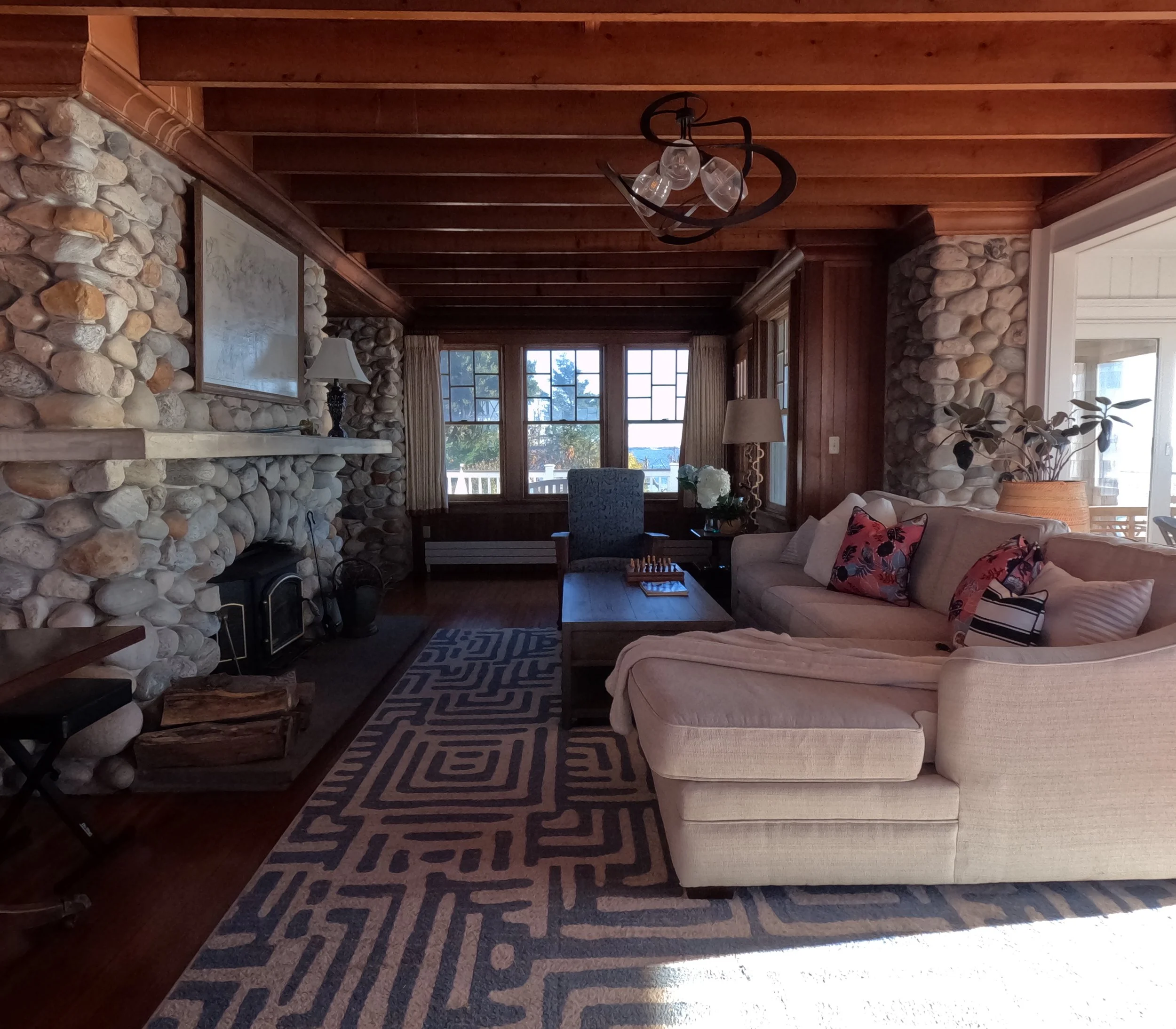 Living room with a stone fireplace, beige sofa with decorative pillows, a wooden coffee table, an armchair, and large windows showing an outdoor view.