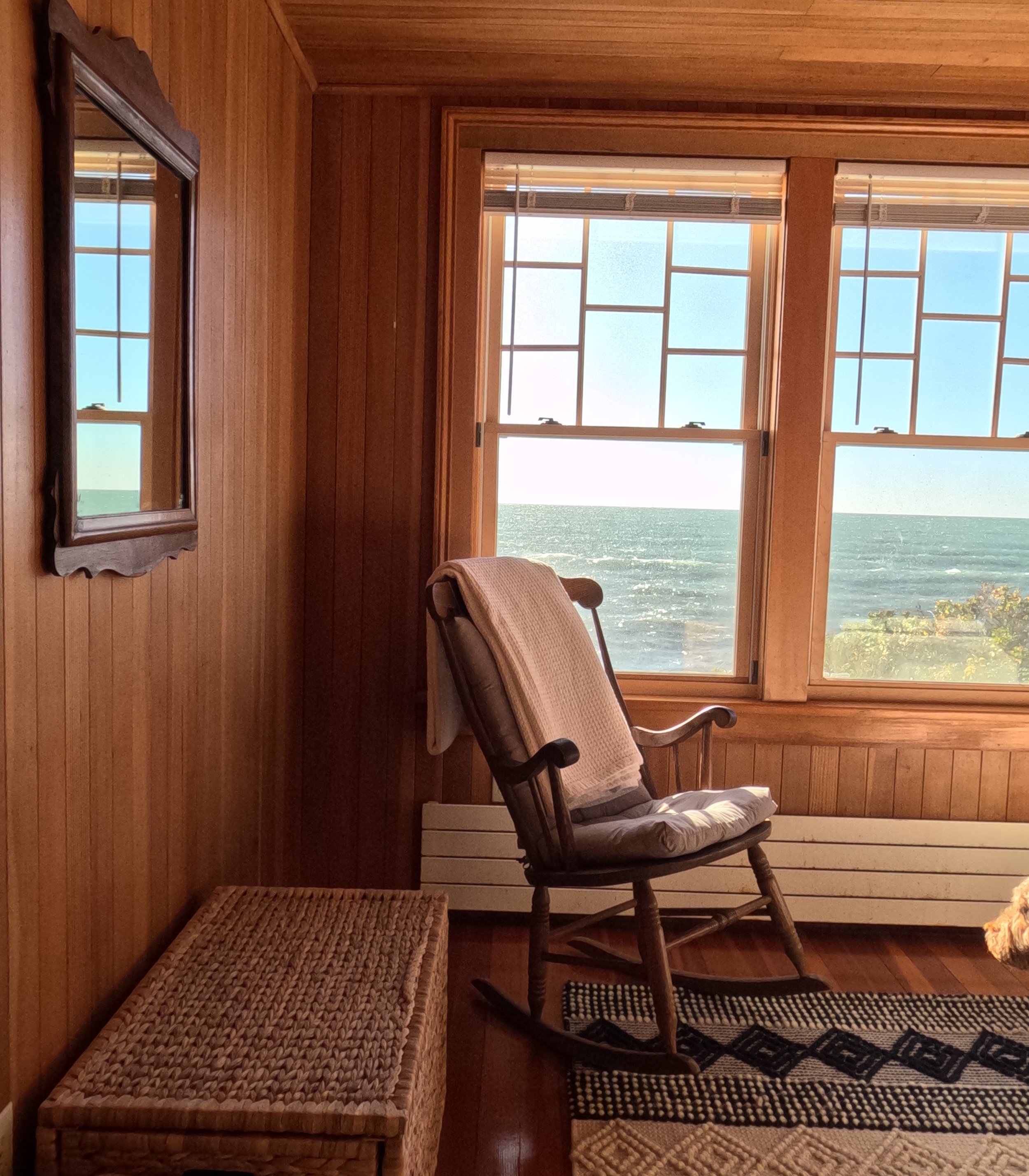A cozy room with wooden walls and large windows showing the ocean outside. There is a rocking chair with a pillow and a blanket, a wicker storage bench, a wall mirror, and a patterned rug.