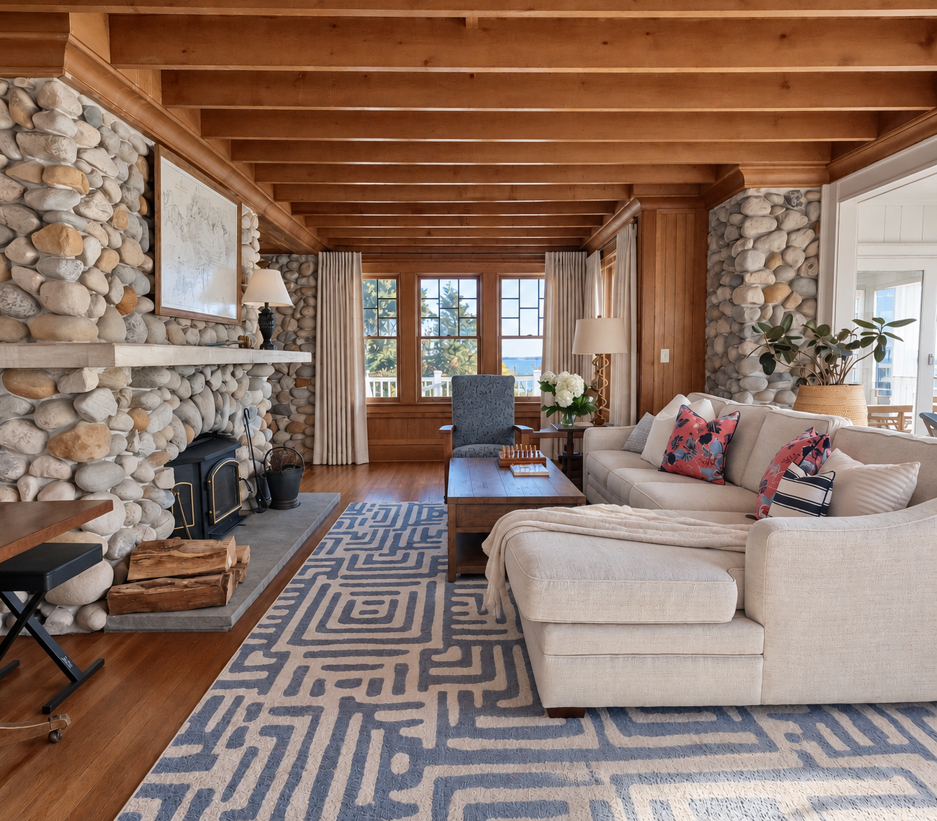 Living room with stone fireplace, beige sectional sofa with colorful pillows, wooden ceiling and walls, large windows with white curtains, coffee table, patterned rug, armchair, side table with lamp, indoor plant, and sliding glass door leading outsi