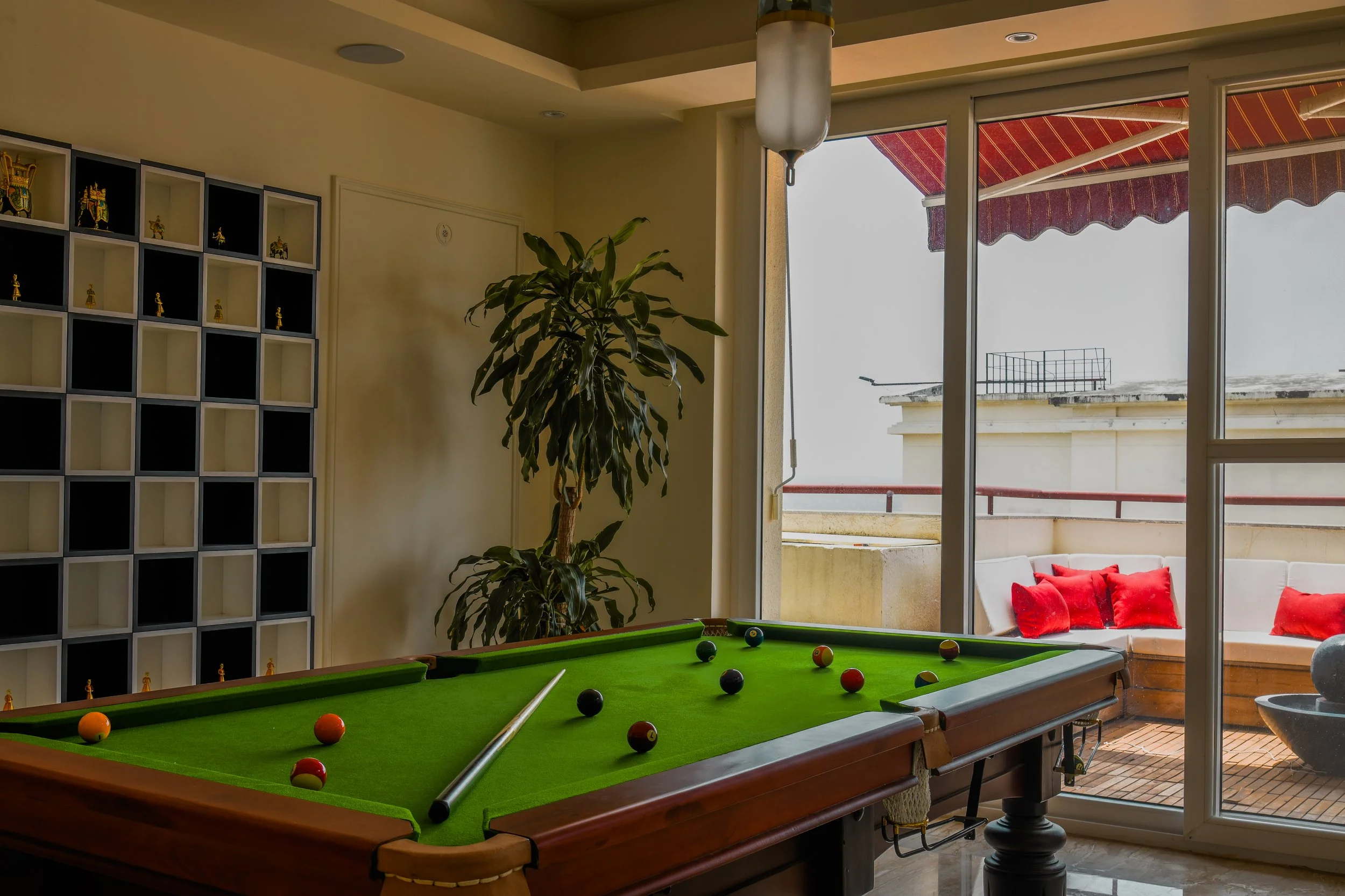 Indoor room with a pool table, a large plant, and a sliding glass door leading to a balcony with outdoor seating and red cushions. Part of a shelving unit with small figurines is visible on the left.