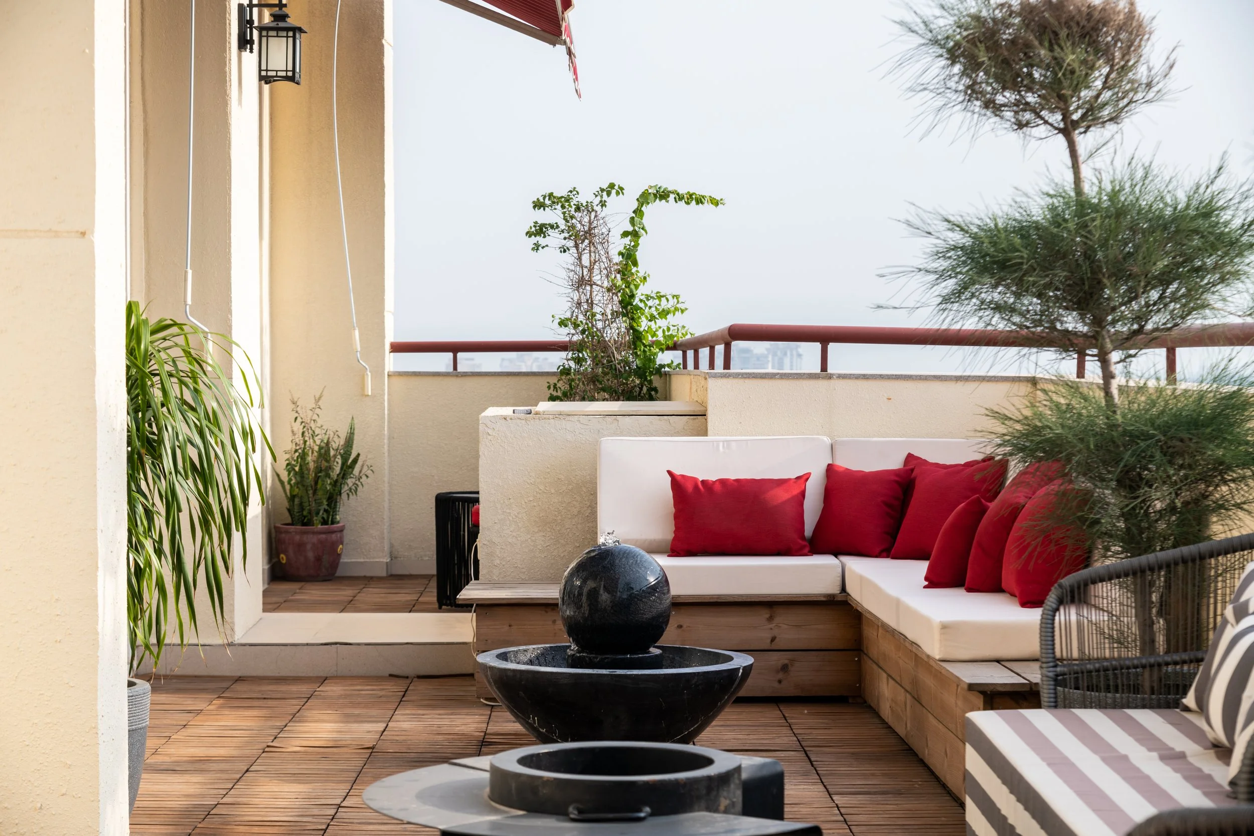 Balcony outdoor seating area with beige walls, red cushions on white benches, potted plants, and a water fountain, with trees and buildings in the background.