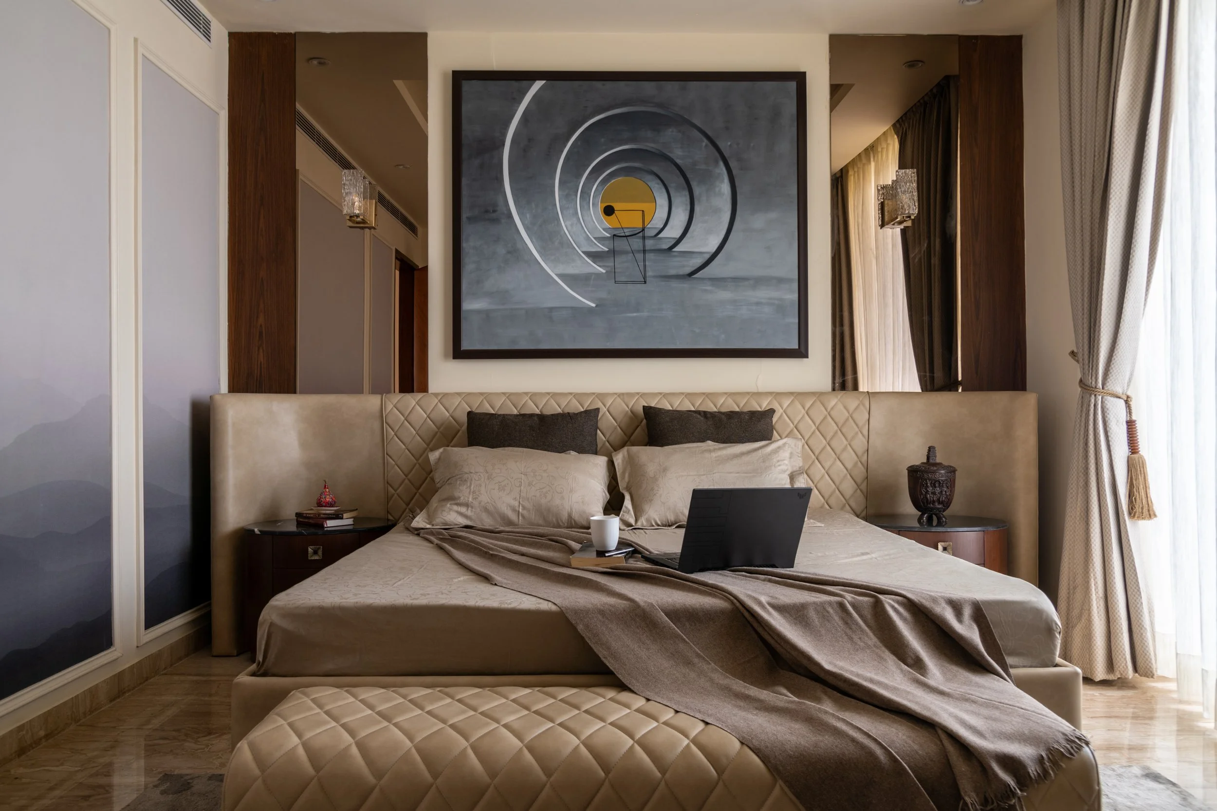 A luxurious bedroom with a beige quilted bed, pillows, a laptop, and a cup on the bed. Two bedside tables with decorative items on either side. Large artwork with concentric circles and geometric shapes hangs above the bed. Curtains are open, allowin
