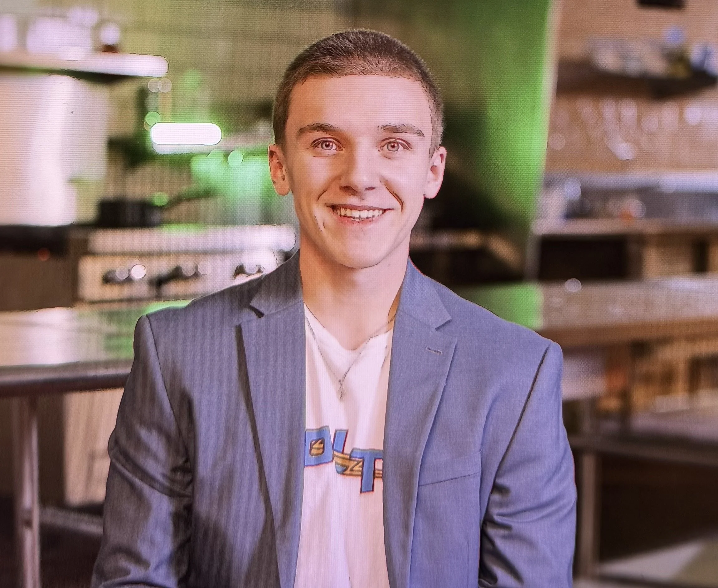 A young man with short hair smiling, wearing a blazer over a graphic T-shirt that says Voltz, sitting in a kitchen.