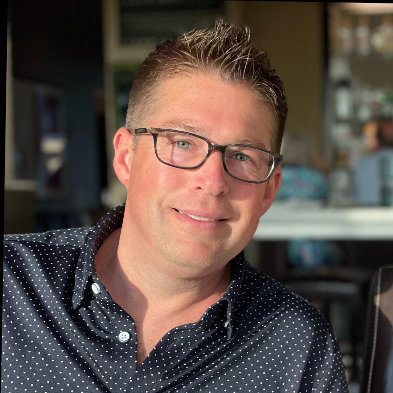A smiling man wearing glasses and a dark, polka-dotted shirt, sitting indoors with a blurred background.