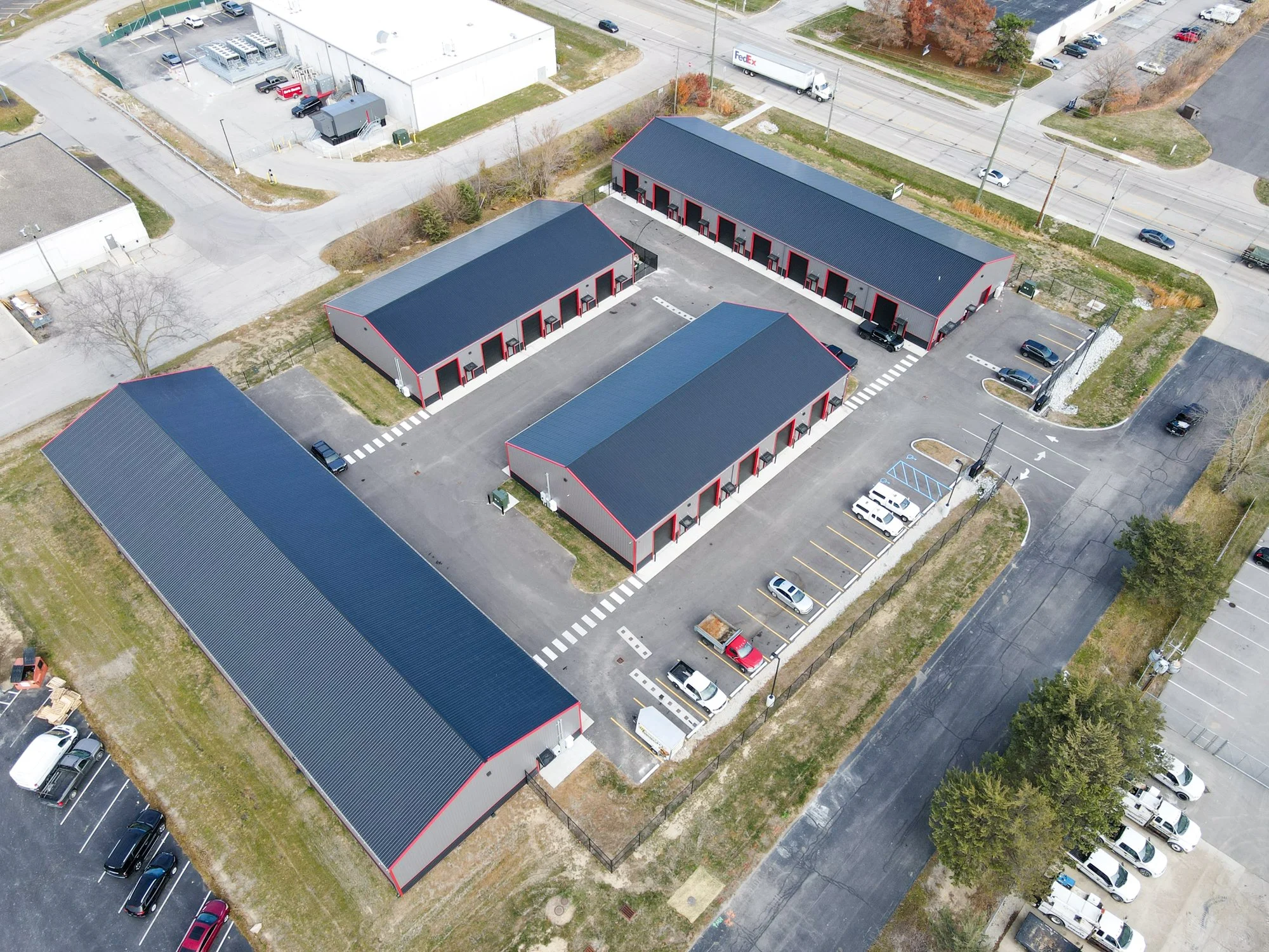 An aerial view of a small commercial building complex with red and gray exteriors, surrounded by parking lots with several parked cars, in an urban area with roads and other buildings nearby.