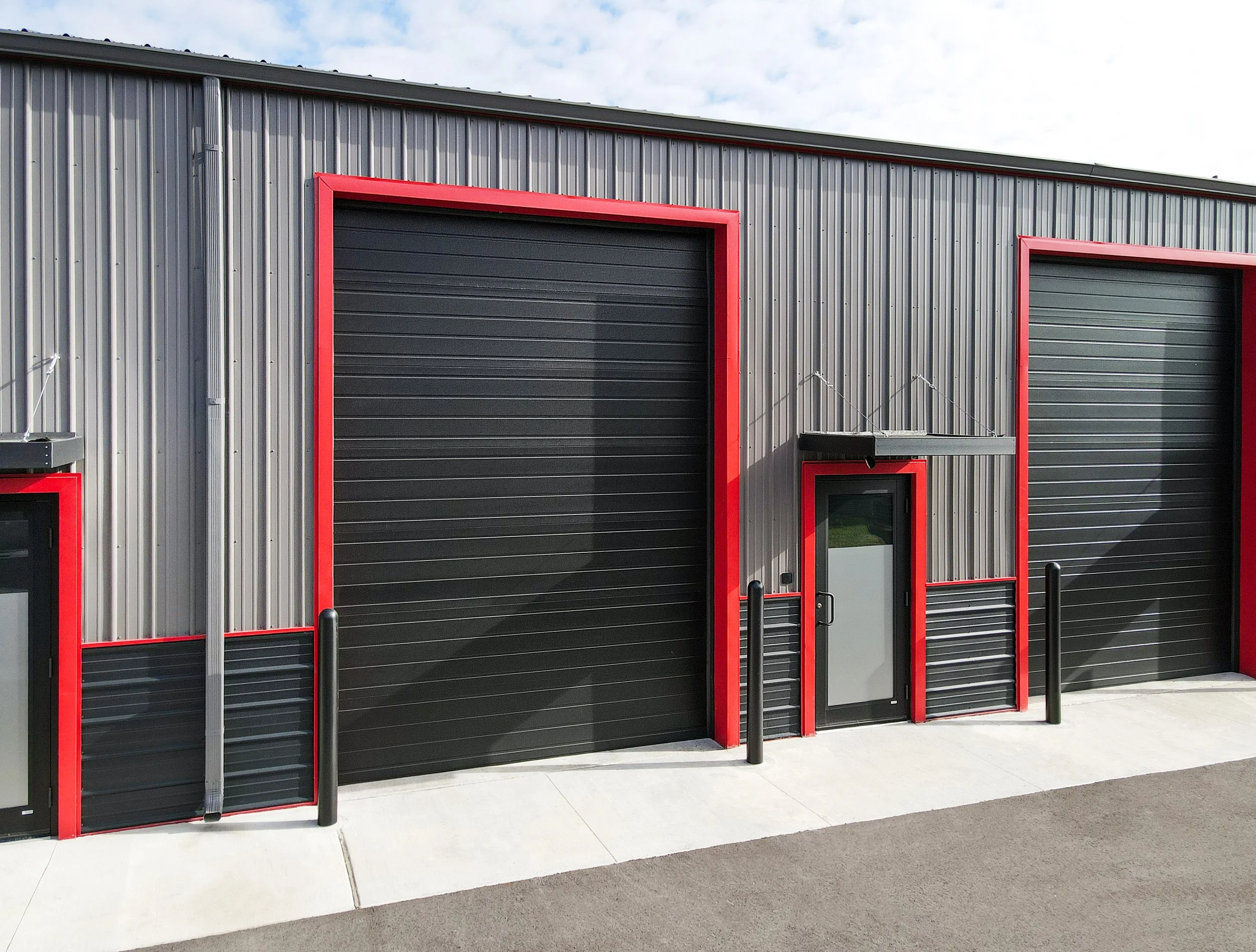 Exterior of a commercial building with three roll-up garage doors, painted black with red framing. The building has gray corrugated metal siding and a sloped roof, with a partly cloudy sky above.
