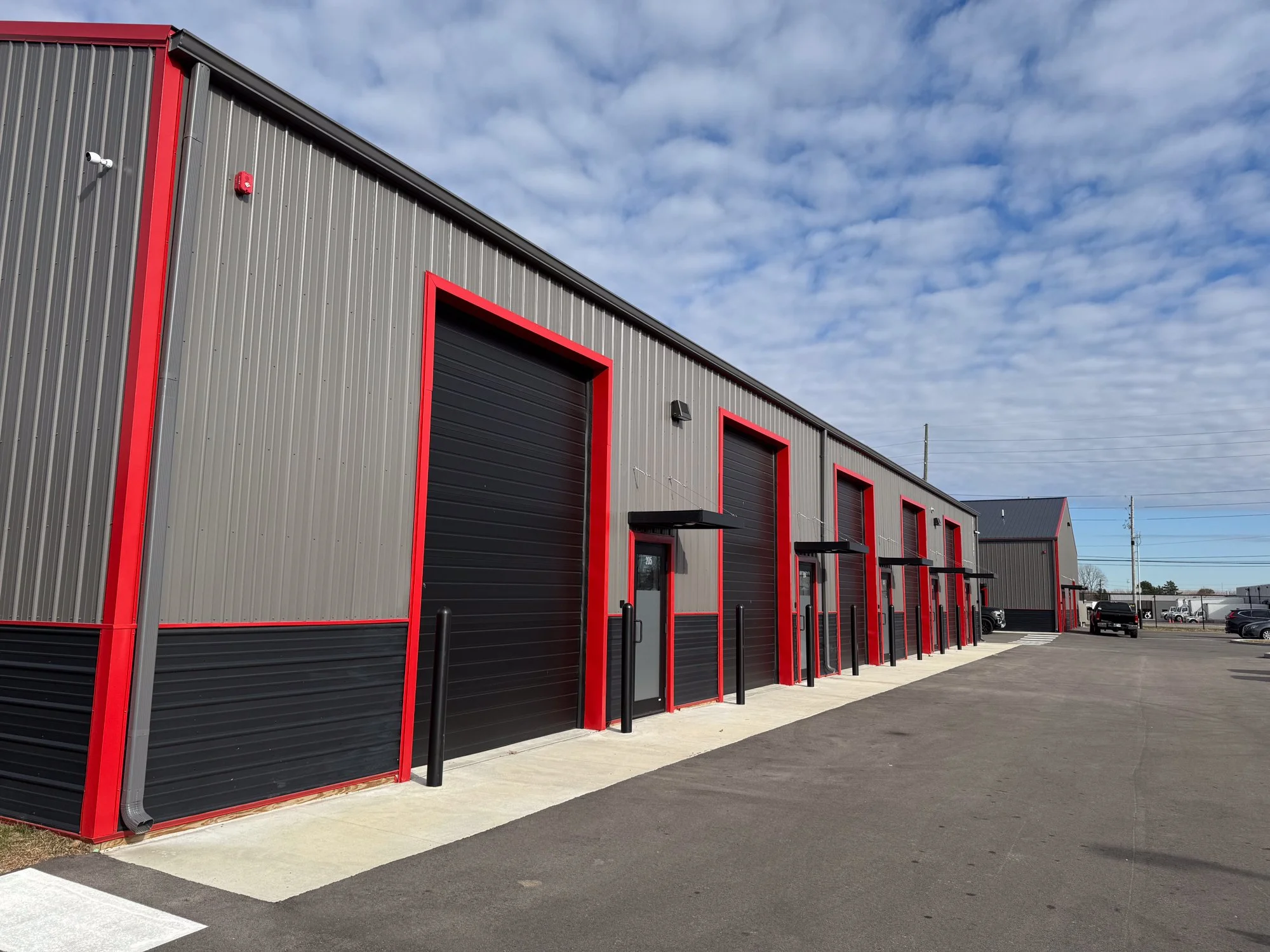 Commercial industrial building with large black garage doors outlined in red and small entrances with black awnings, in a parking lot with a few parked cars, under a partly cloudy sky.