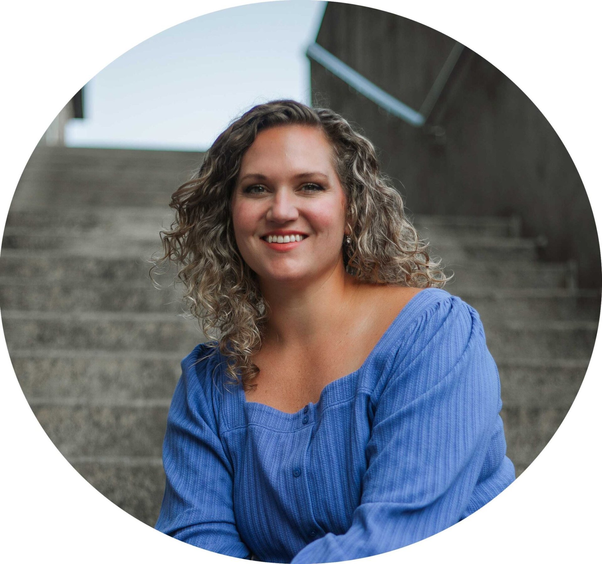 A woman with shoulder-length curly hair, wearing a blue top, smiles at the camera while sitting on outdoor steps.