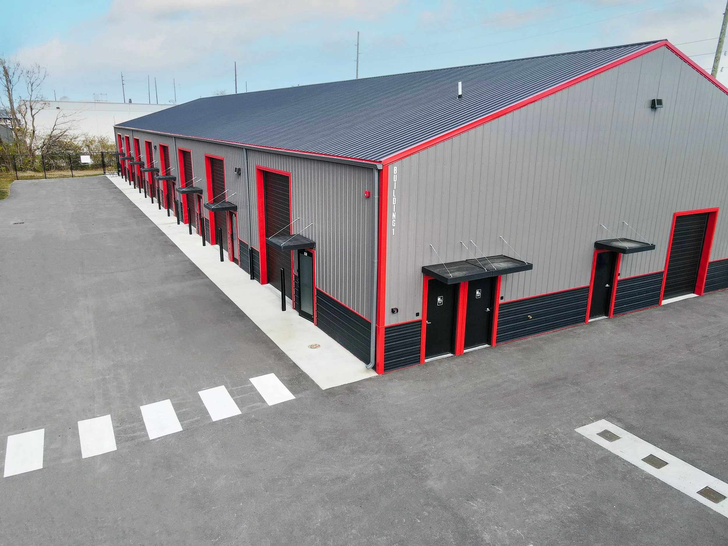 Large industrial building with a metal exterior, gray with red accents, multiple garage doors, and loading docks, situated on an asphalt lot with a crosswalk and trees in the background.