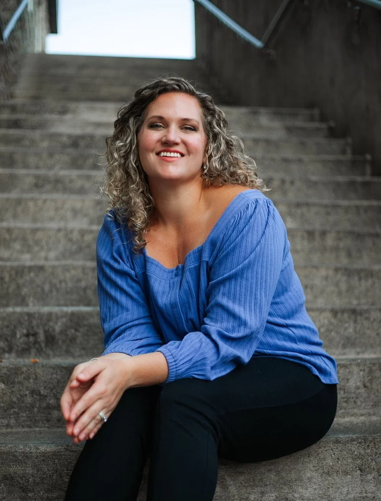 A woman with curly blonde hair smiling, sitting on outdoor concrete stairs, wearing a blue long-sleeve top and black pants.