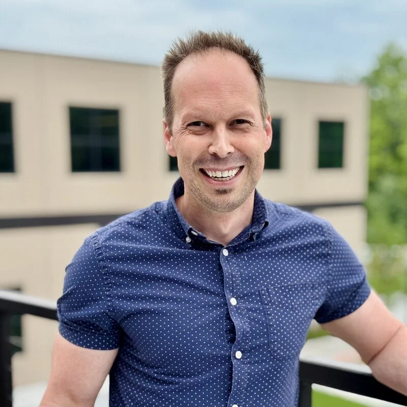 A smiling man with short hair wearing a blue polka dot shirt, outdoors near a building with large windows, during daytime.