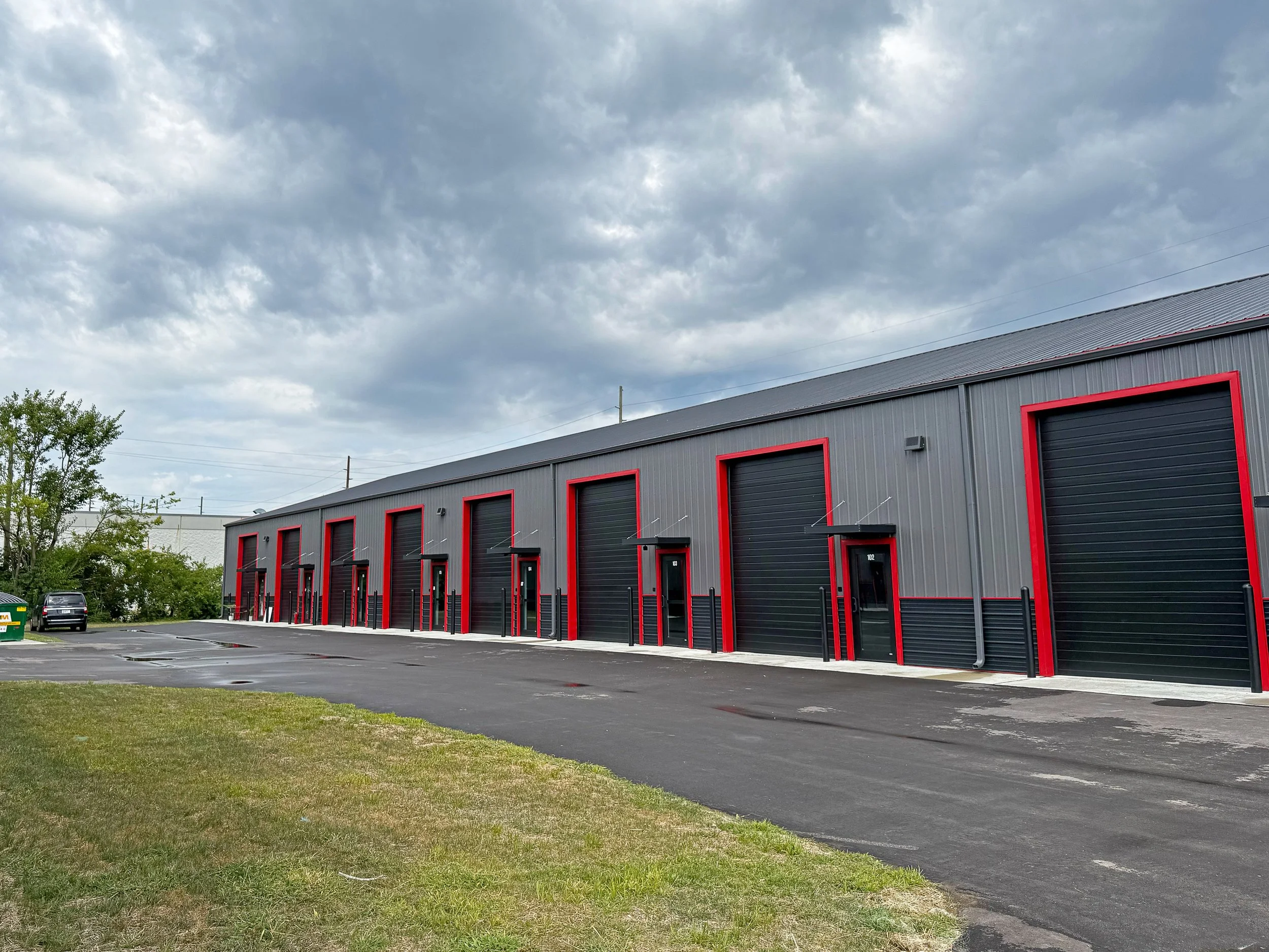A commercial warehouse with black garage doors and red framing, located in an outdoor area with a paved driveway and a grassy lawn in the foreground. Cars are parked to the left, and stormy clouds are present in the sky.