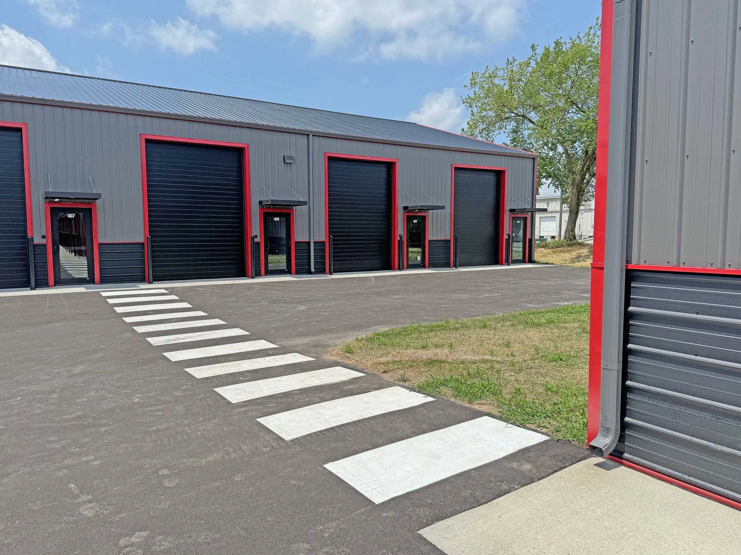 Outside of a commercial storage facility with multiple black roll-up garage doors, each framed with red trim, and pedestrian doors. A crosswalk is painted on the asphalt in front of the buildings with a grassy area and trees in the background under a partly cloudy sky.