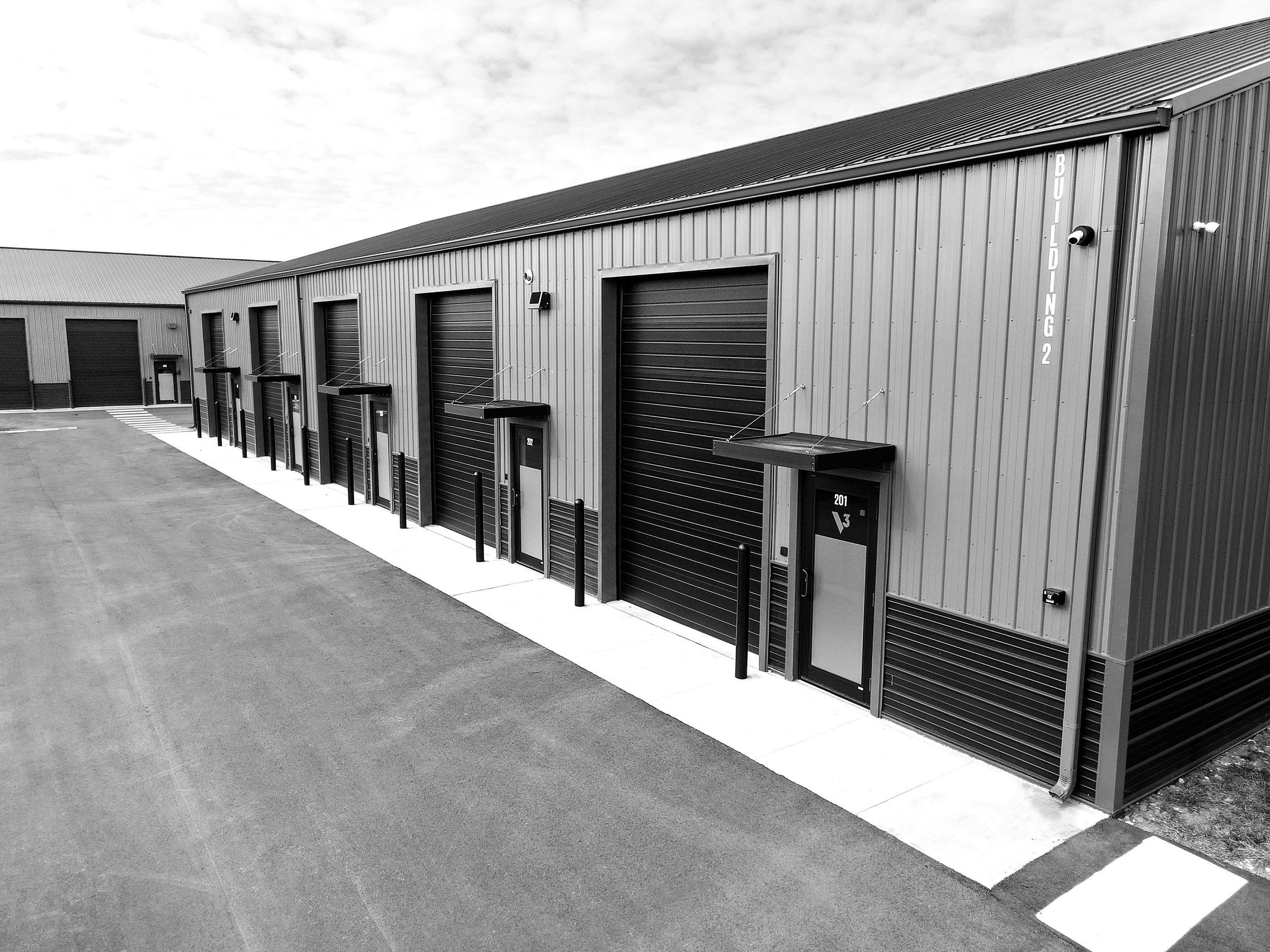 Row of industrial storage units with roll-up doors and small entrances, surrounded by an empty paved area.