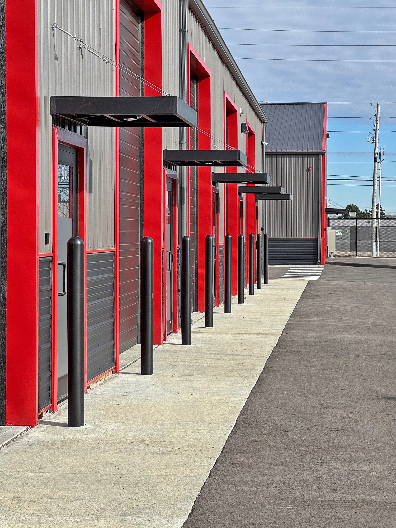 Exterior view of a commercial building with gray and red siding, black awnings above doors, and black safety bollards along the sidewalk, under a partly cloudy sky.