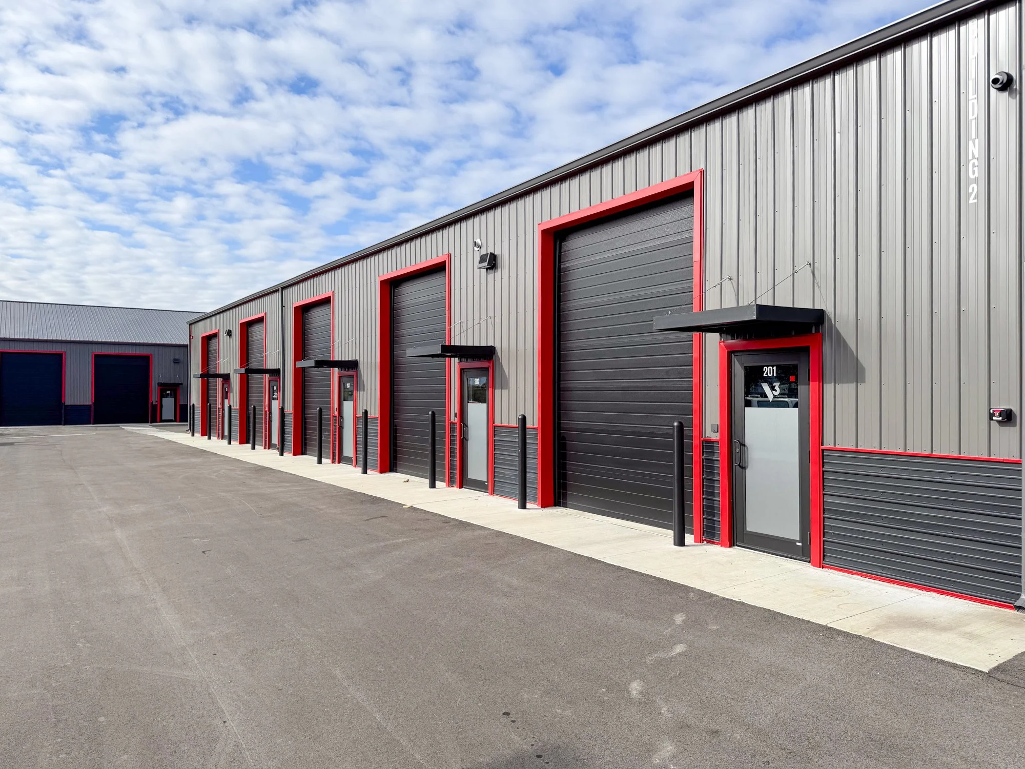 A row of industrial warehouse units with black roll-up doors framed in red, small pedestrian doors, and outdoor service lights, under a sky with scattered clouds.