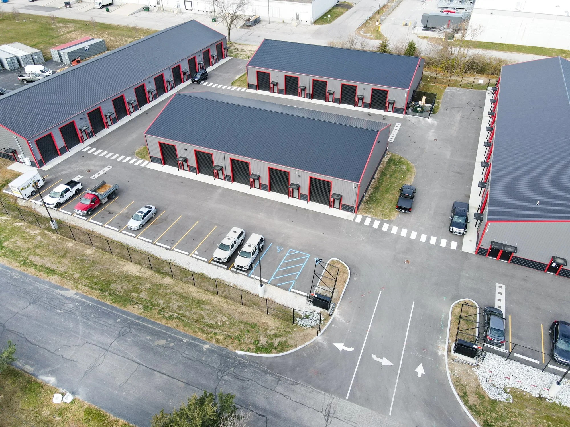 Aerial view of a commercial warehouse complex with multiple grey buildings with red accents, surrounded by parking lots with cars and trucks, and roadways with directional arrows.