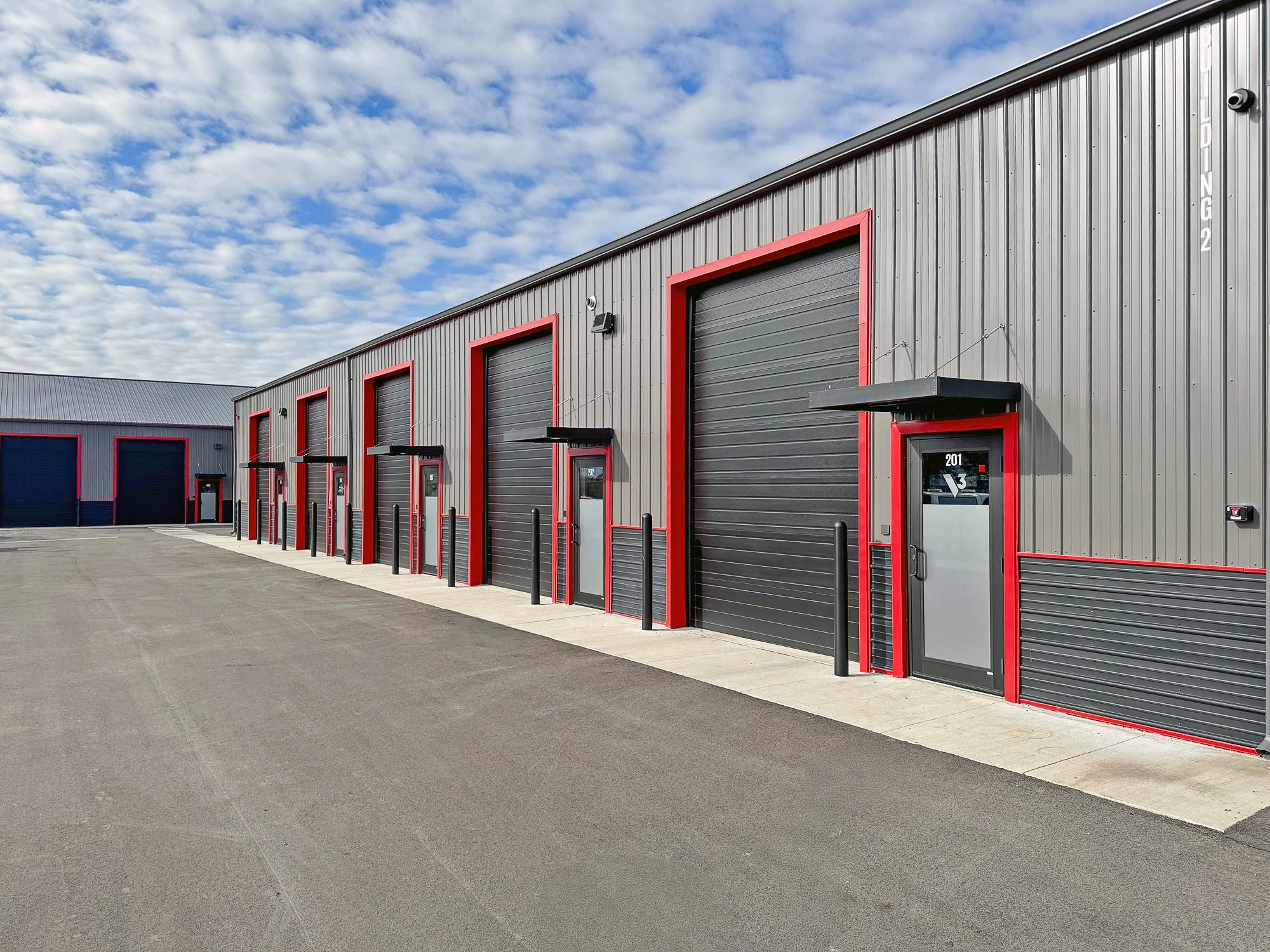 A row of industrial storage units with black roll-up doors, red trim, and black bollards outside, under a partly cloudy sky.