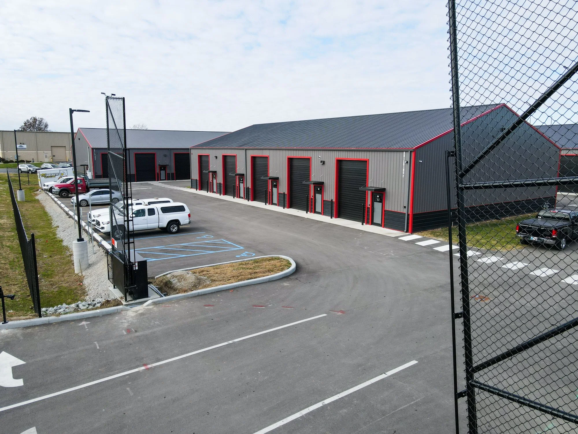 A commercial warehouse with red and black trim, multiple loading bays, parking lot with vehicles, including disabled spaces, surrounded by a chain-link fence.