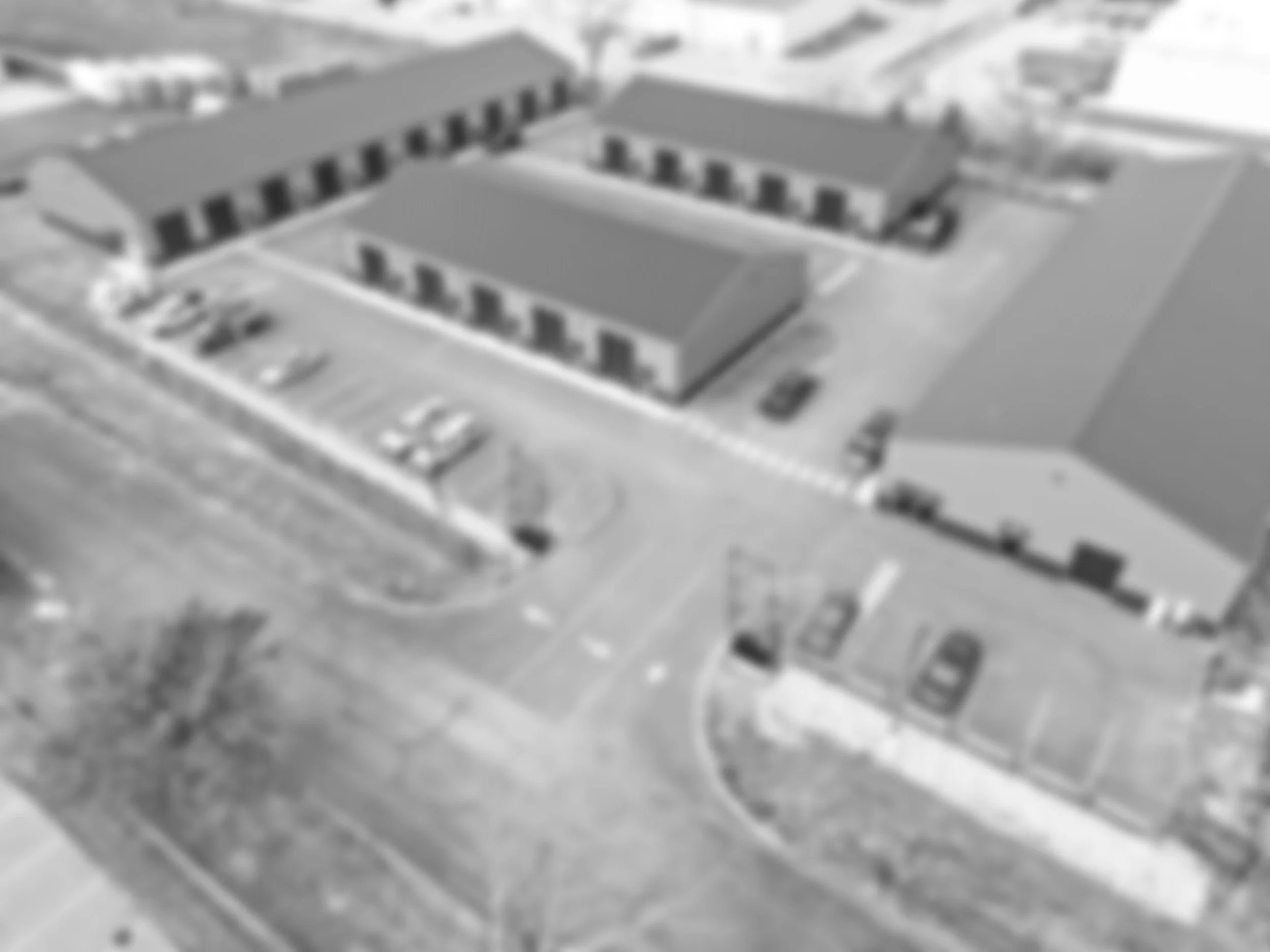 A grayscale aerial view of a building complex with multiple sections, parking lots, driveways, and surrounding roads.