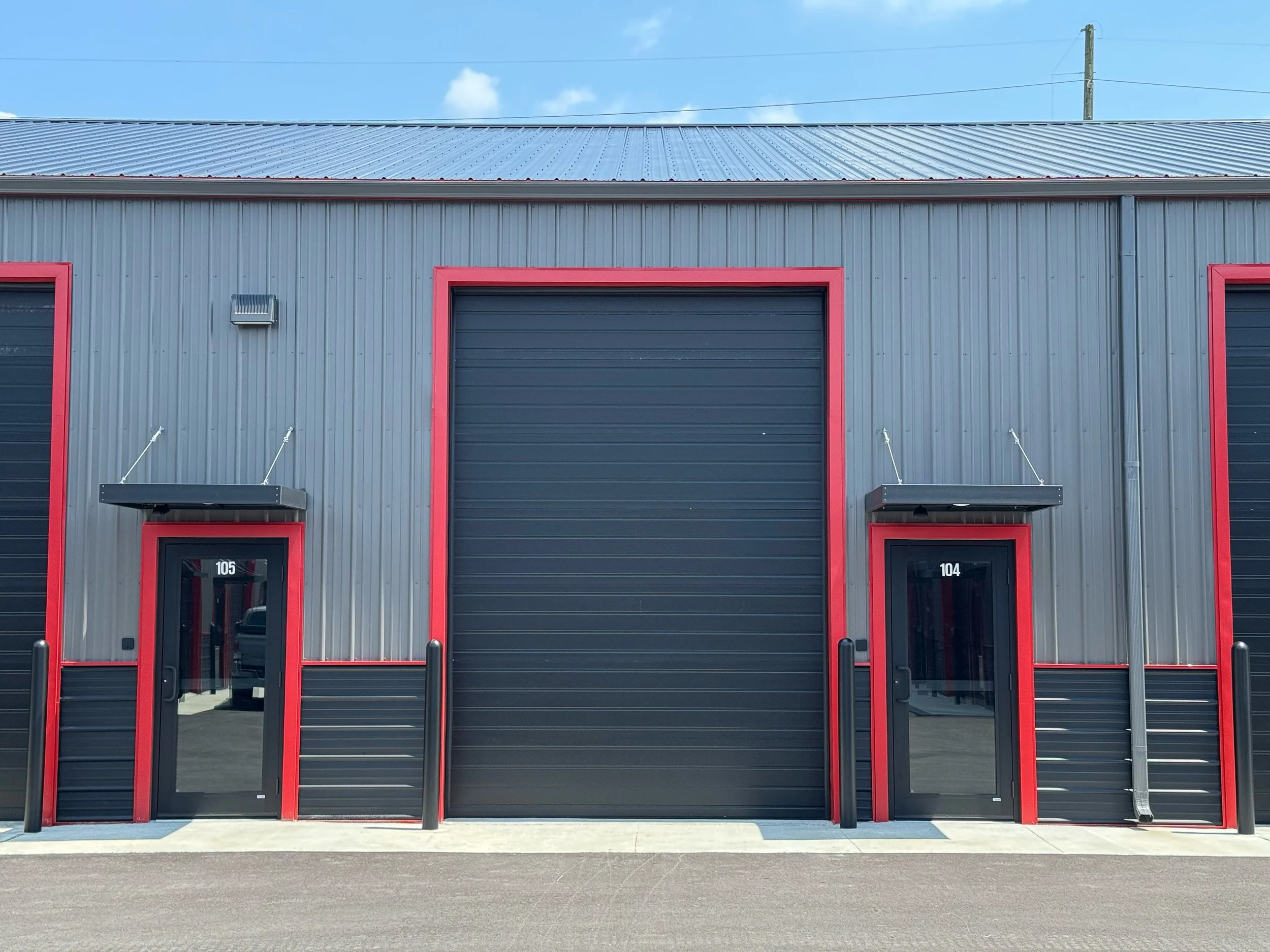 Exterior of a gray industrial warehouse building with red trim, black roll-up door, and two office doors with glass panels, numbered 105 and 104.