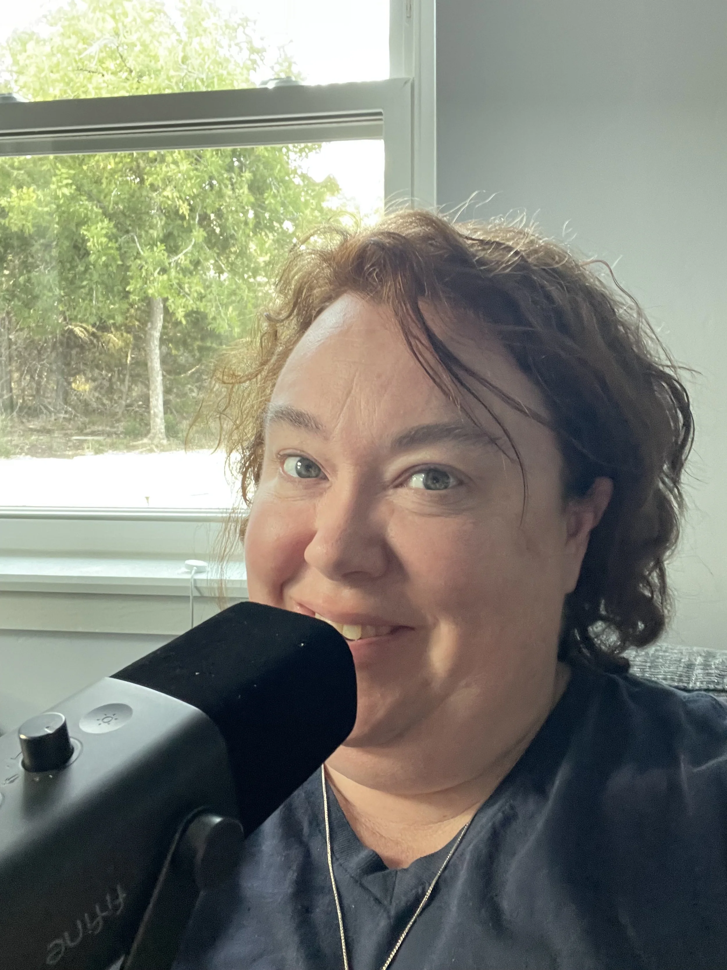 A person with curly brown hair smiling and speaking into a black microphone in front of a window with green trees outside.