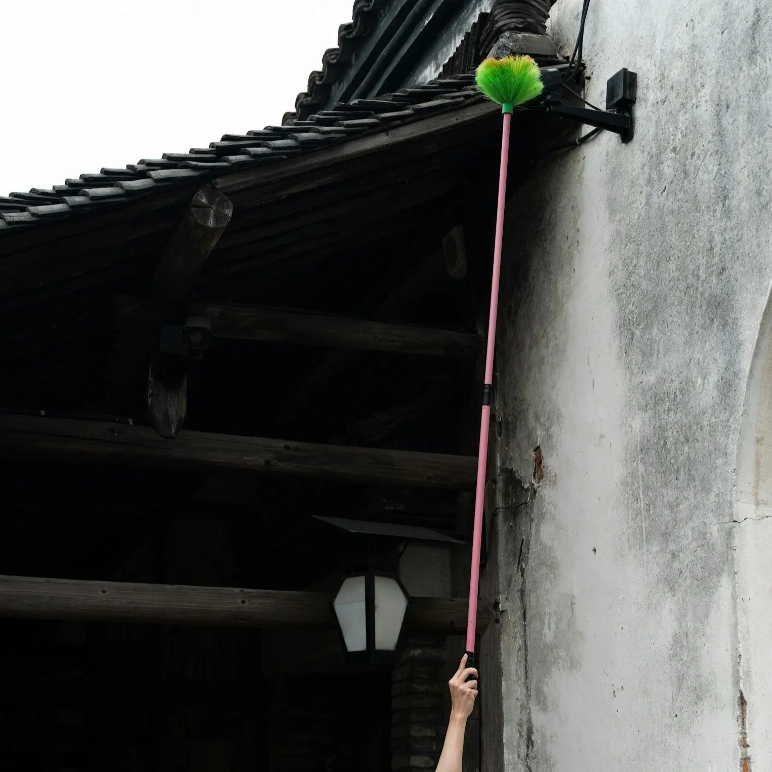 A person is holding a long pink pole with a green duster at the end, cleaning the corner of a building's wall under the roof.