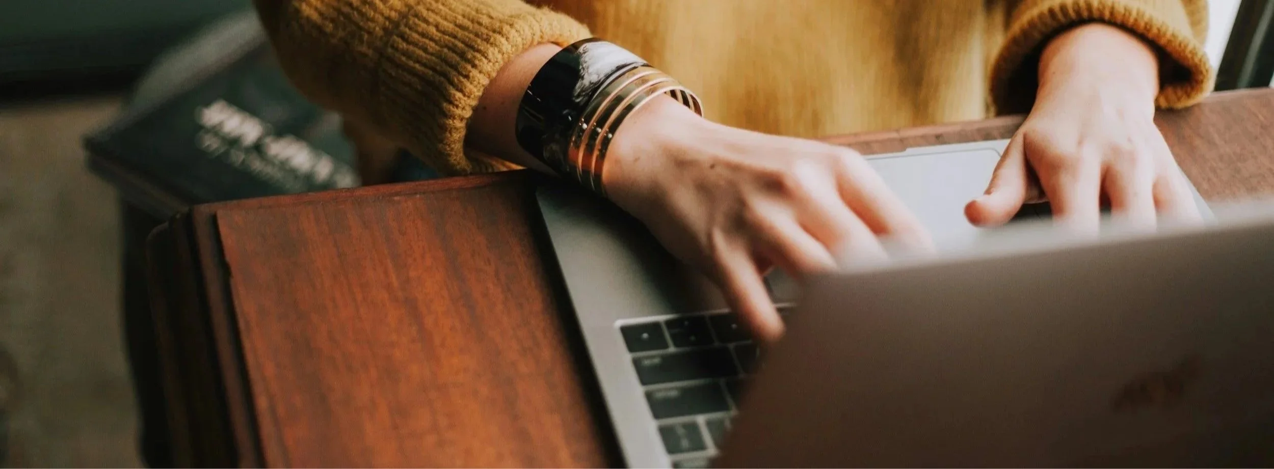 A person wearing a mustard-colored sweater and a chunky bracelet is typing on a silver laptop with a wooden desk background.