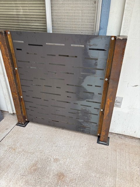 Metal decorative panel with horizontal slits, framed by rust-colored metal posts, placed outdoors on concrete ground.