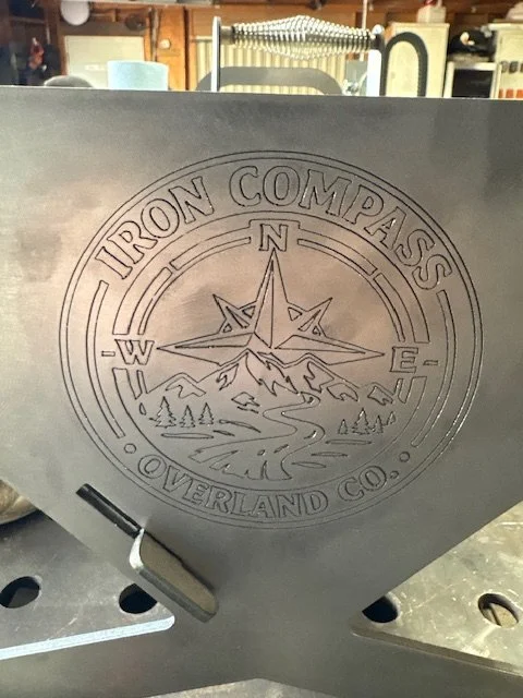 Metal sheet with the engraved logo of Iron Compass, a company based in Overland, Co., featuring a compass rose, mountains, trees, and a winding river.
