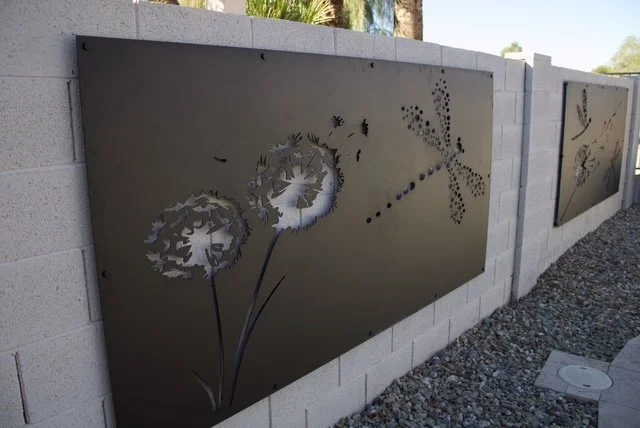 Metal art panels mounted on a white brick wall featuring a dandelion and a dragonfly design, with a gravel ground and desert plants in the background.