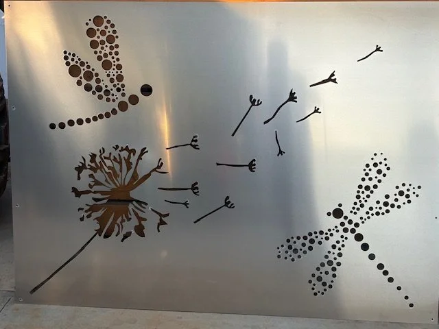 Decorative metal panel with silhouettes of a dragonfly, a dandelion, and a group of birds flying.