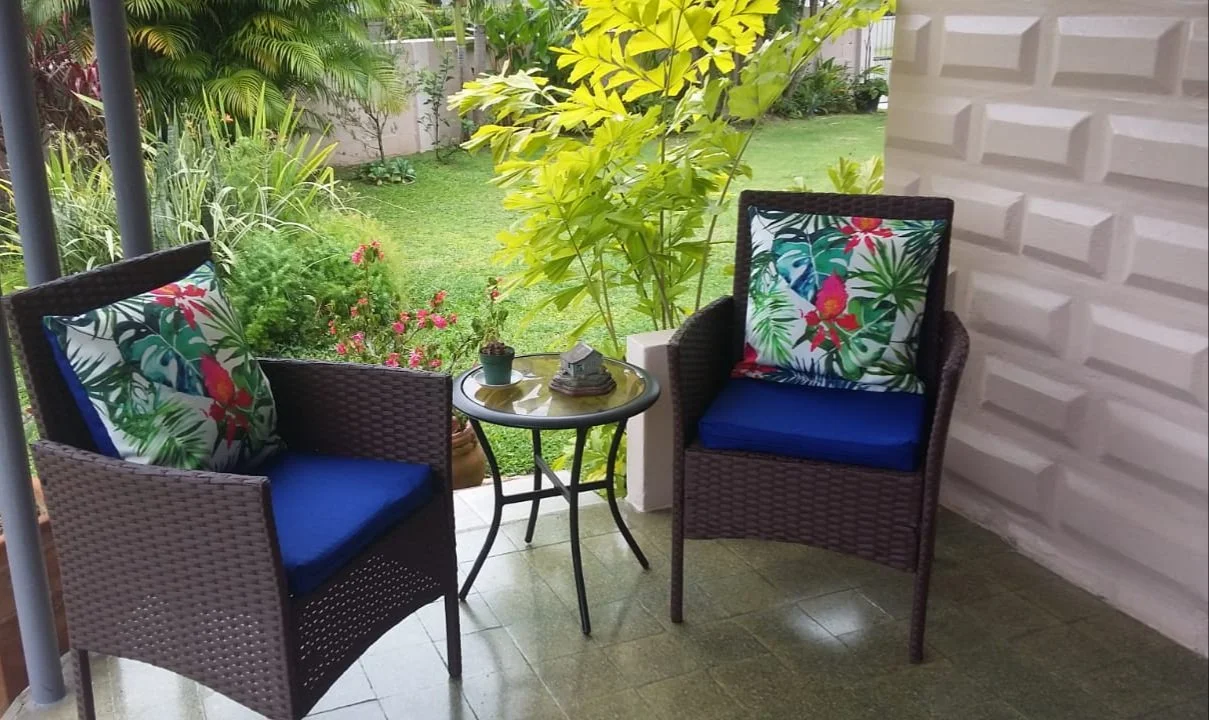 Patio area with two wicker chairs with tropical print cushions, a small round table with a potted plant and a magazine, beside a white textured wall and green garden with plants and trees.