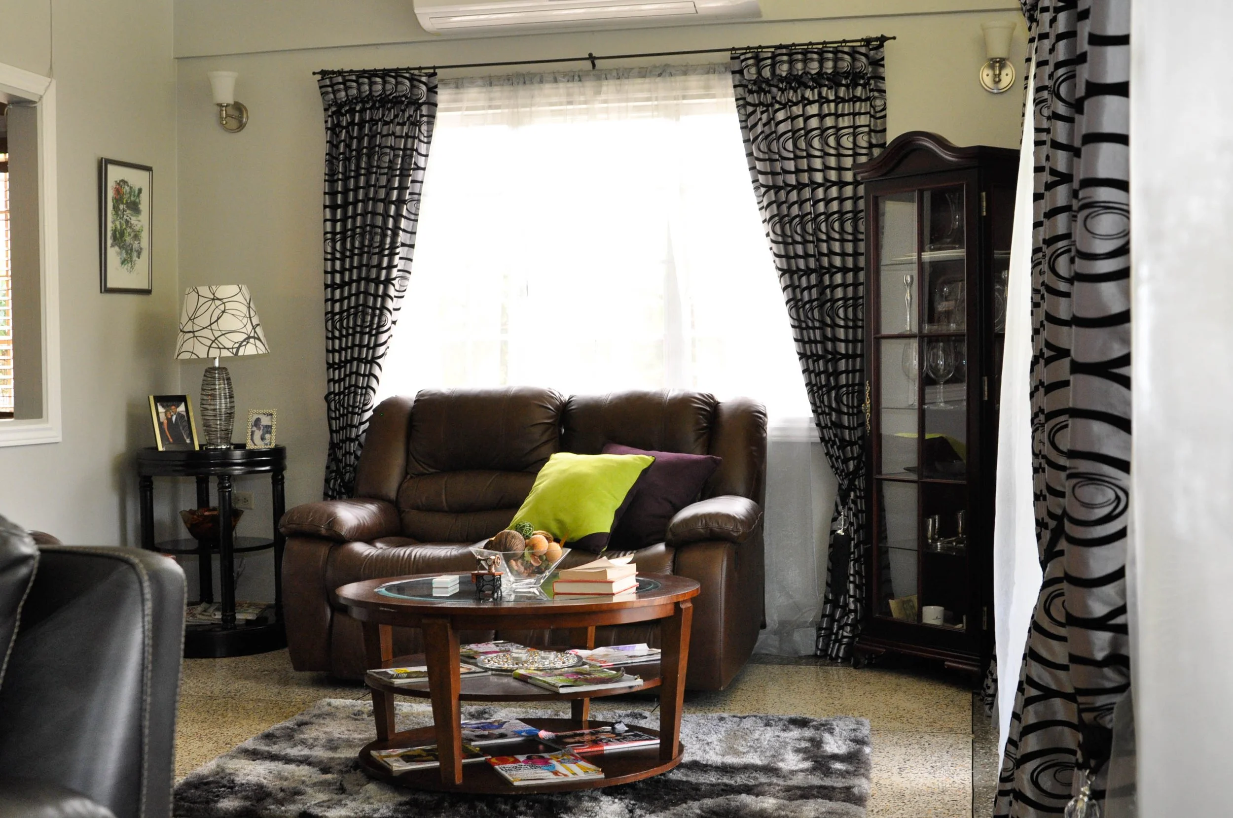 Living room with brown leather sofa, wooden coffee table with magazines, black and white patterned curtains, and a glass-fronted display cabinet. There is a window behind the sofa with sheer white curtains, and a side table with photo frames and a la