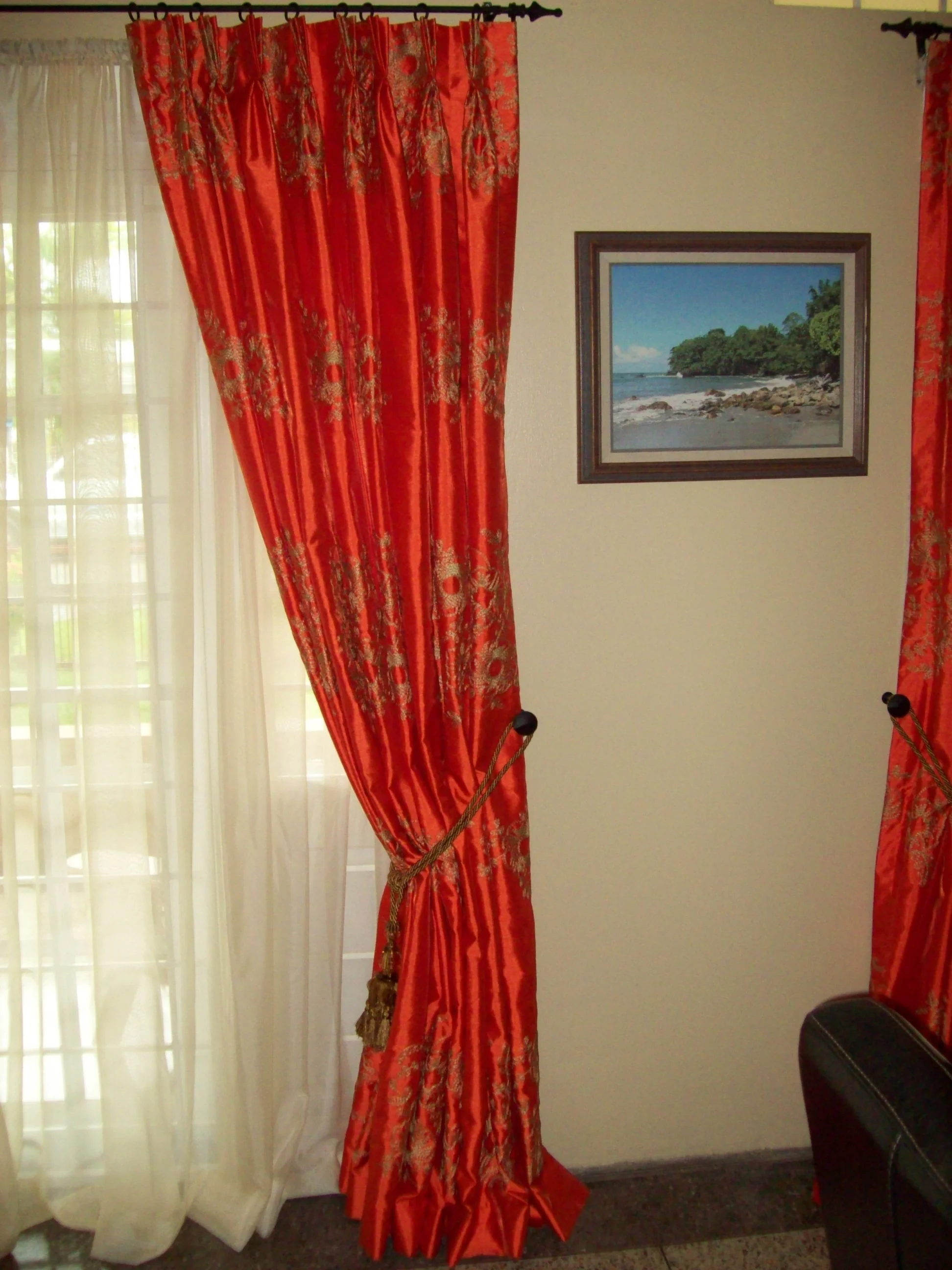 Red curtains with intricate designs tied back revealing a sheer white curtain and a window, with a framed picture of a beach scene on the wall